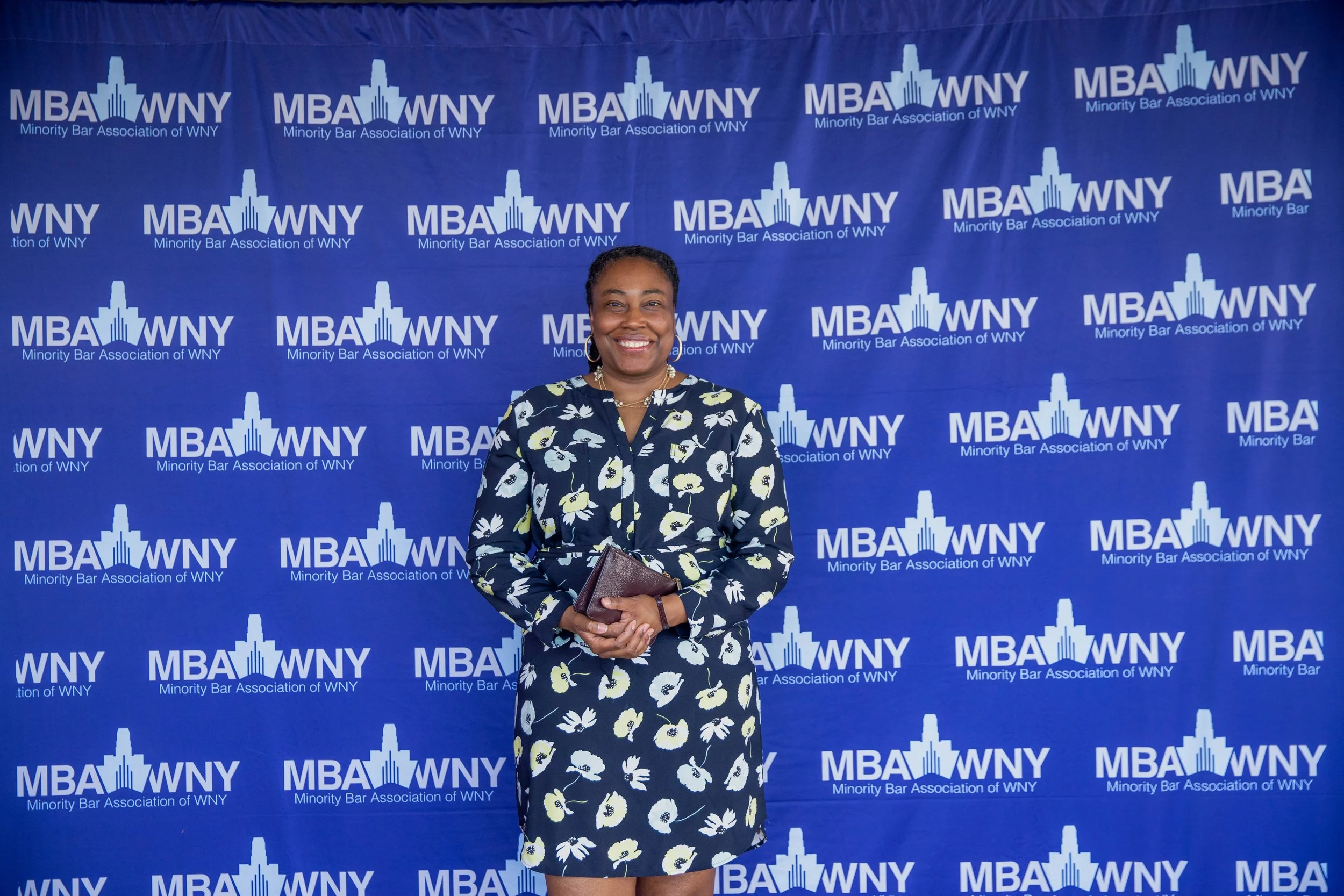 Woman in a floral dress smiling and holding a clutch bag standing in front of a blue backdrop with the logo and text for the Minority Bar Association of WNY.