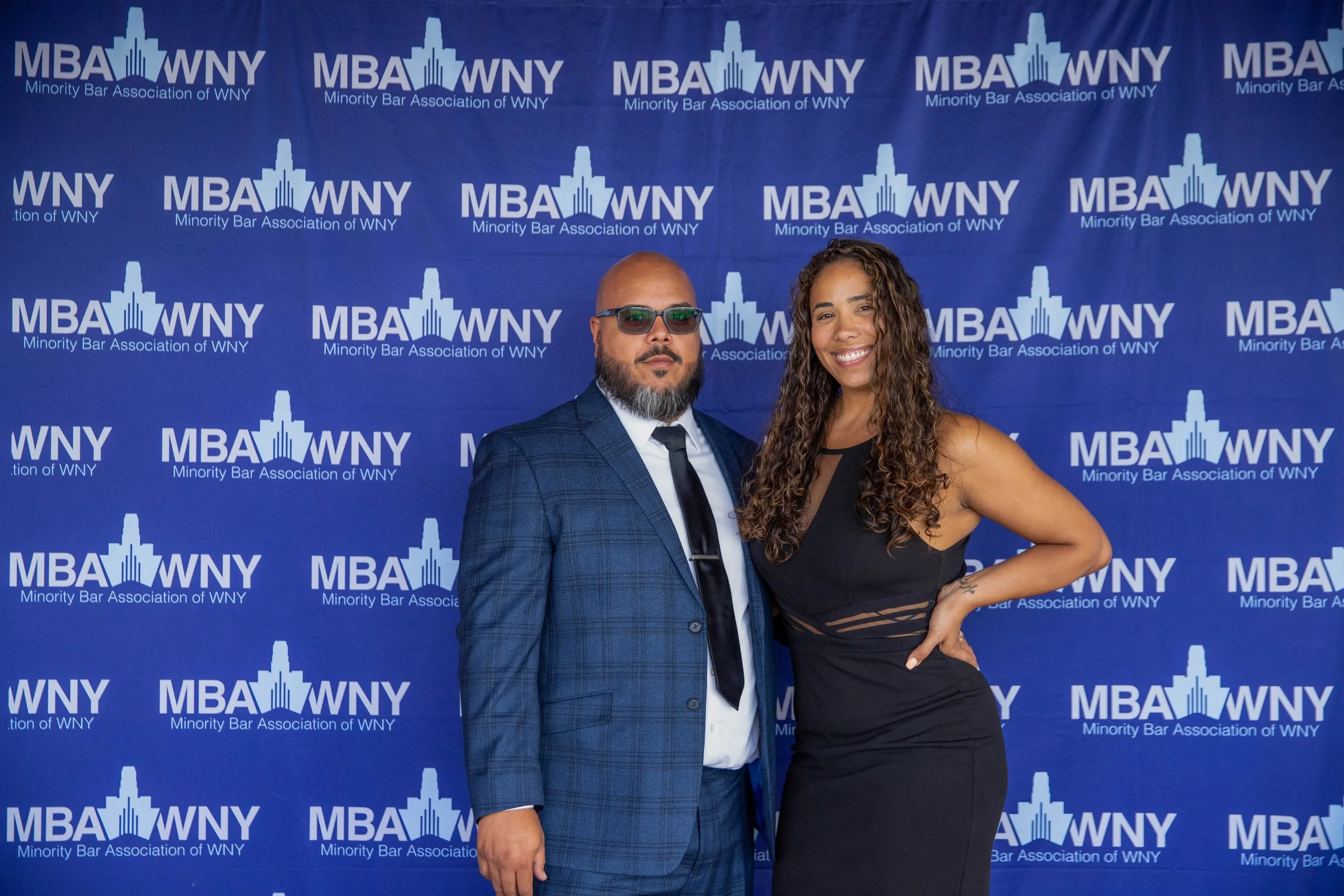 Two people posing in front of a blue backdrop with the logo and text for the Minority Bar Association of WNY. The man on the left is wearing glasses, a blue checkered suit, a white shirt, and a black tie. The woman on the right has long curly hair, i