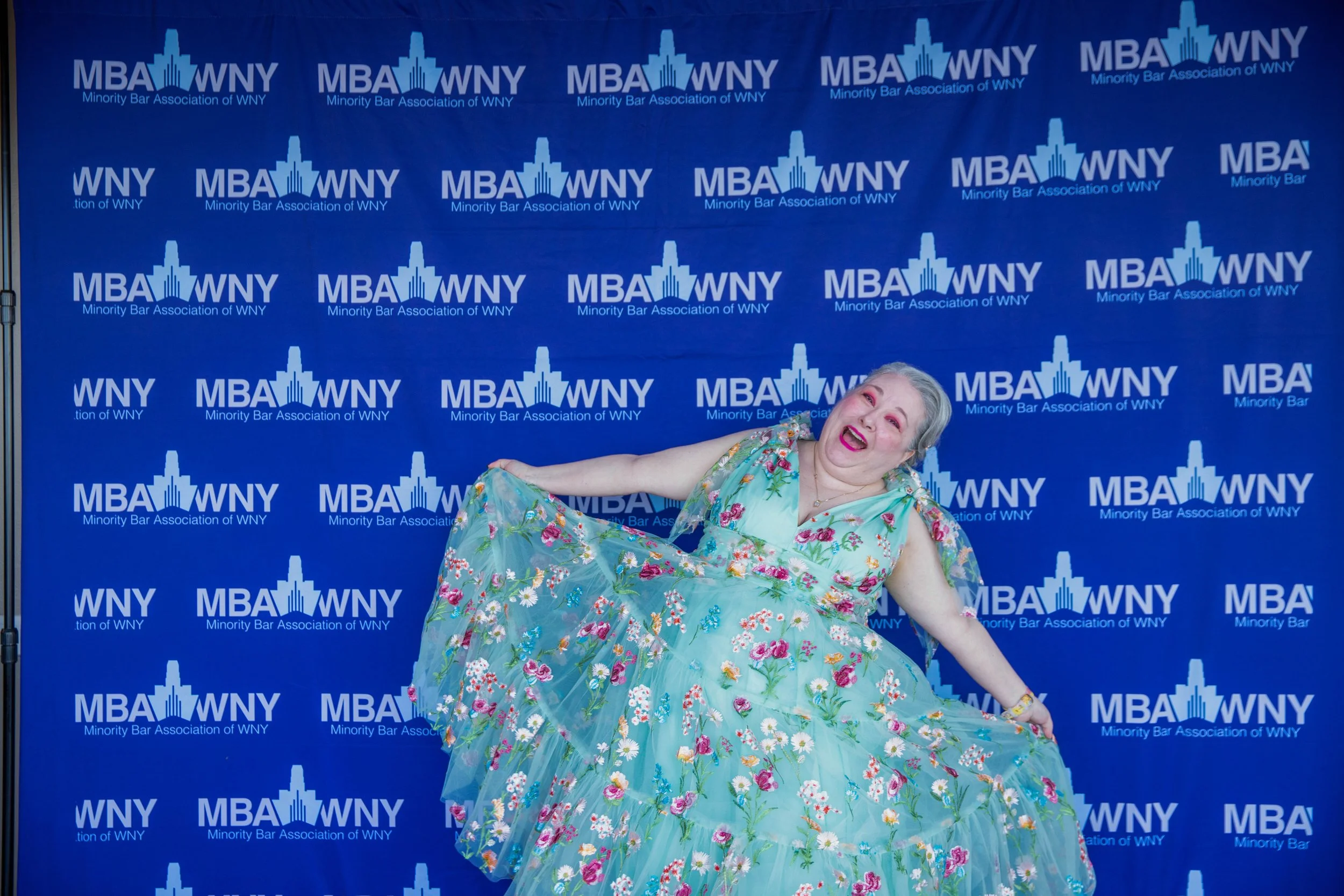 An elderly woman with gray hair and bright pink lipstick wearing a flowing light blue dress with a floral pattern, smiling and posing playfully in front of a blue backdrop with 'MBA WNY Minority Bar Association of WNY' logos.