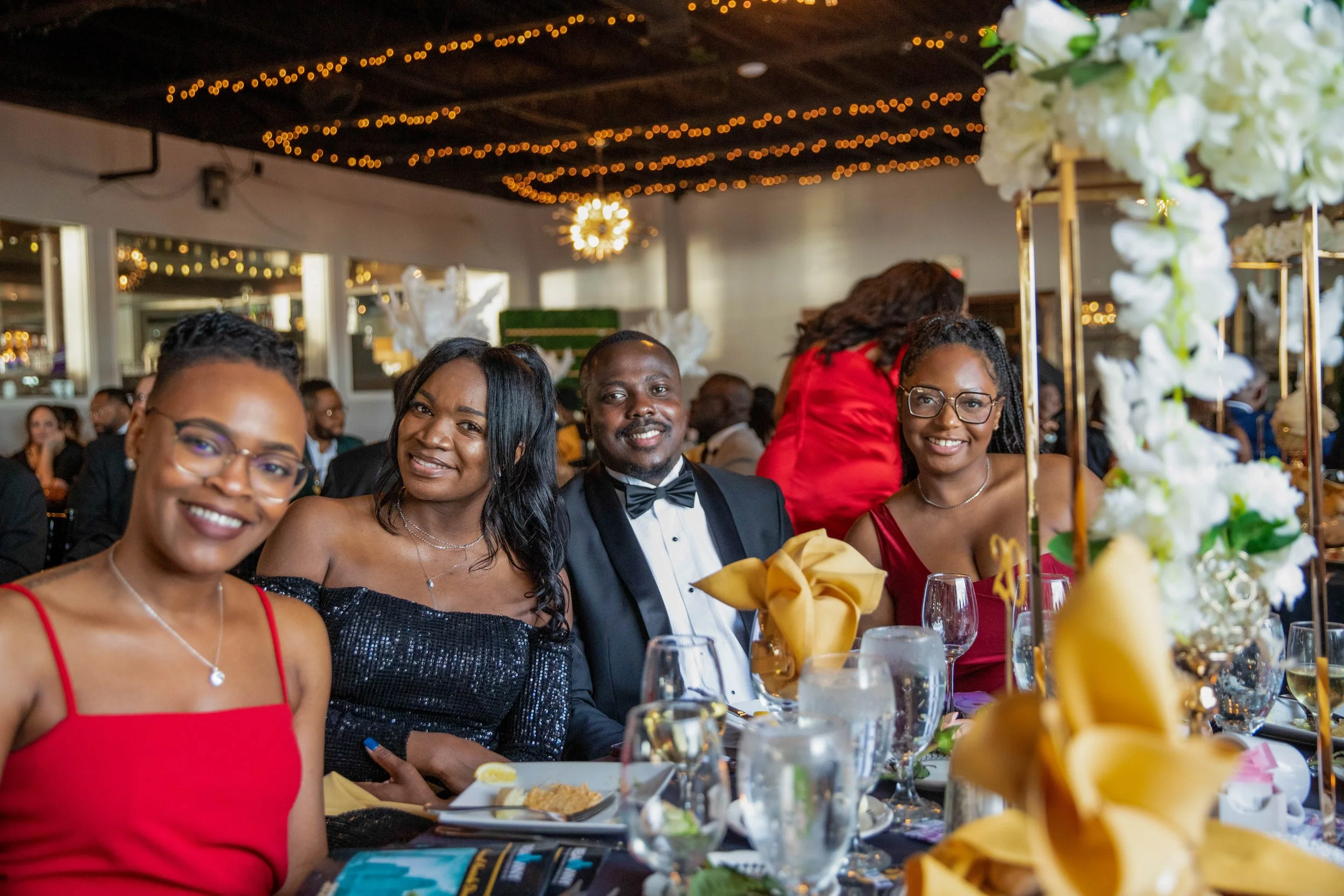 People dressed in formal attire enjoying a celebration at a decorated event venue with string lights and floral centerpieces.