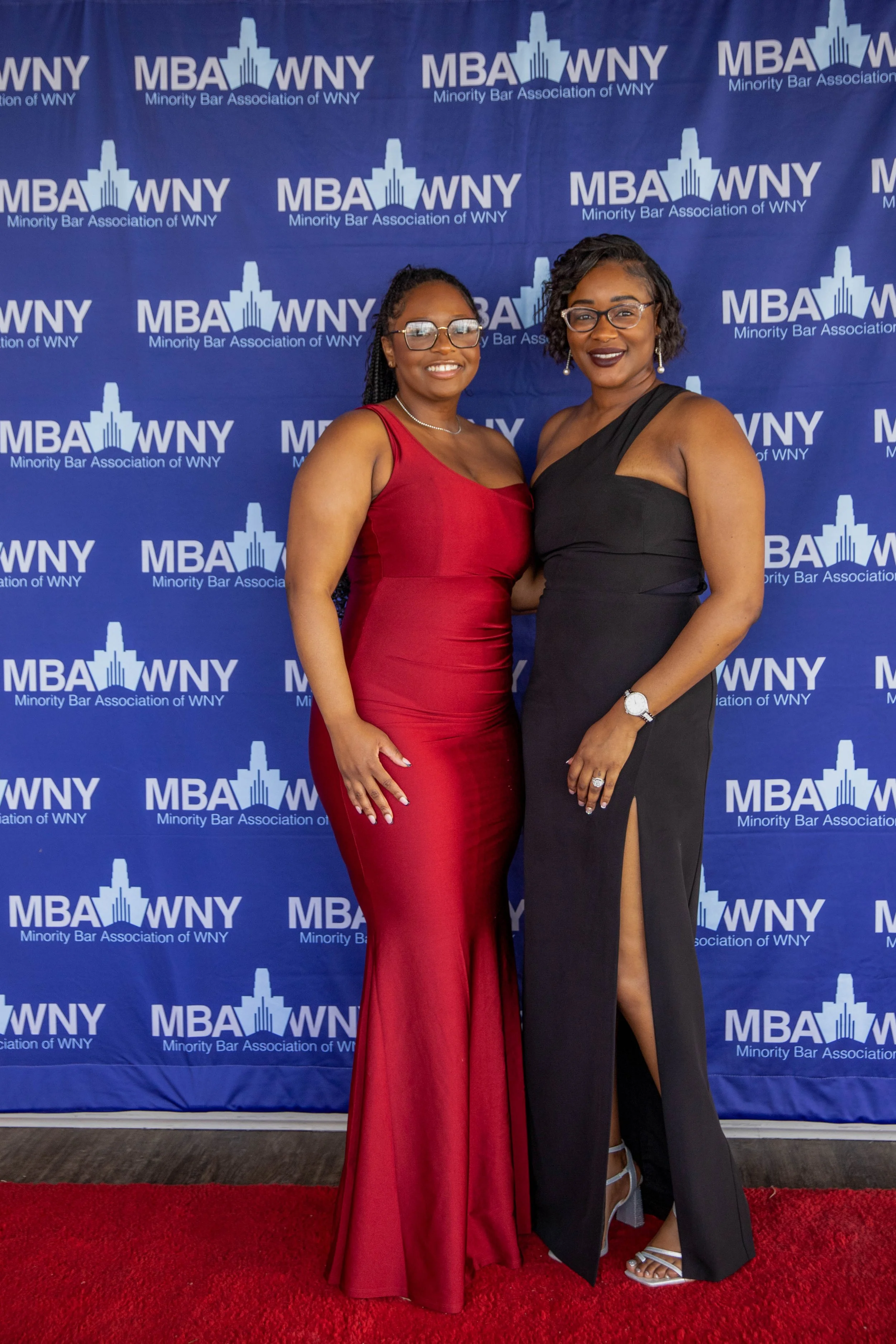 Two women in formal dresses posing together at an event with a blue backdrop that reads 'Minority Bar Association of WNY' and features a logo of a building with a skyscraper silhouette. One woman is wearing a red dress and glasses, and the other is i
