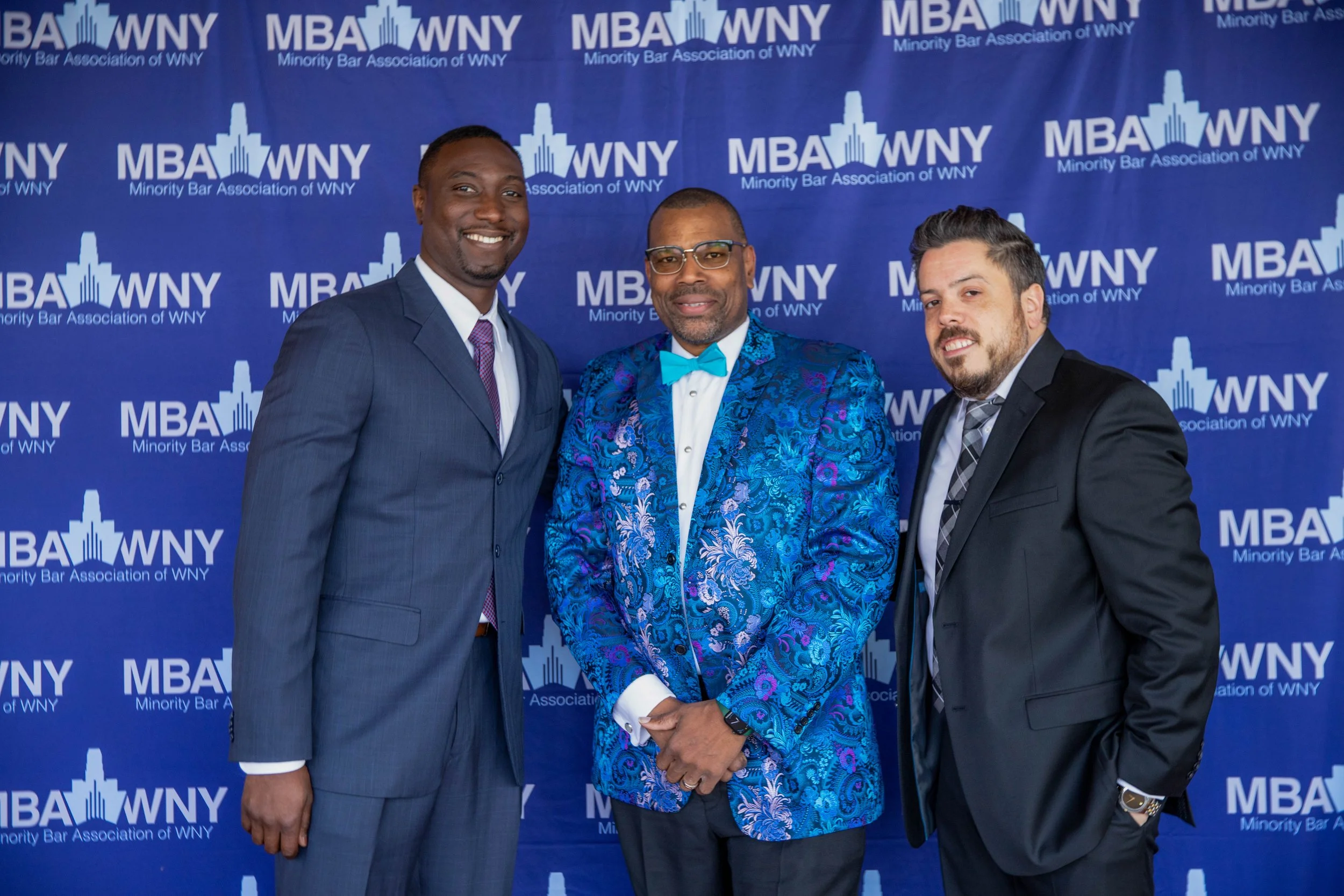 Three men dressed in formal suits standing in front of a blue backdrop with the logo and text 'MBA WNY Minority Bar Association of WNY'. One man wears a dark gray suit with a purple tie, another wears a bright blue patterned suit with a matching bow 