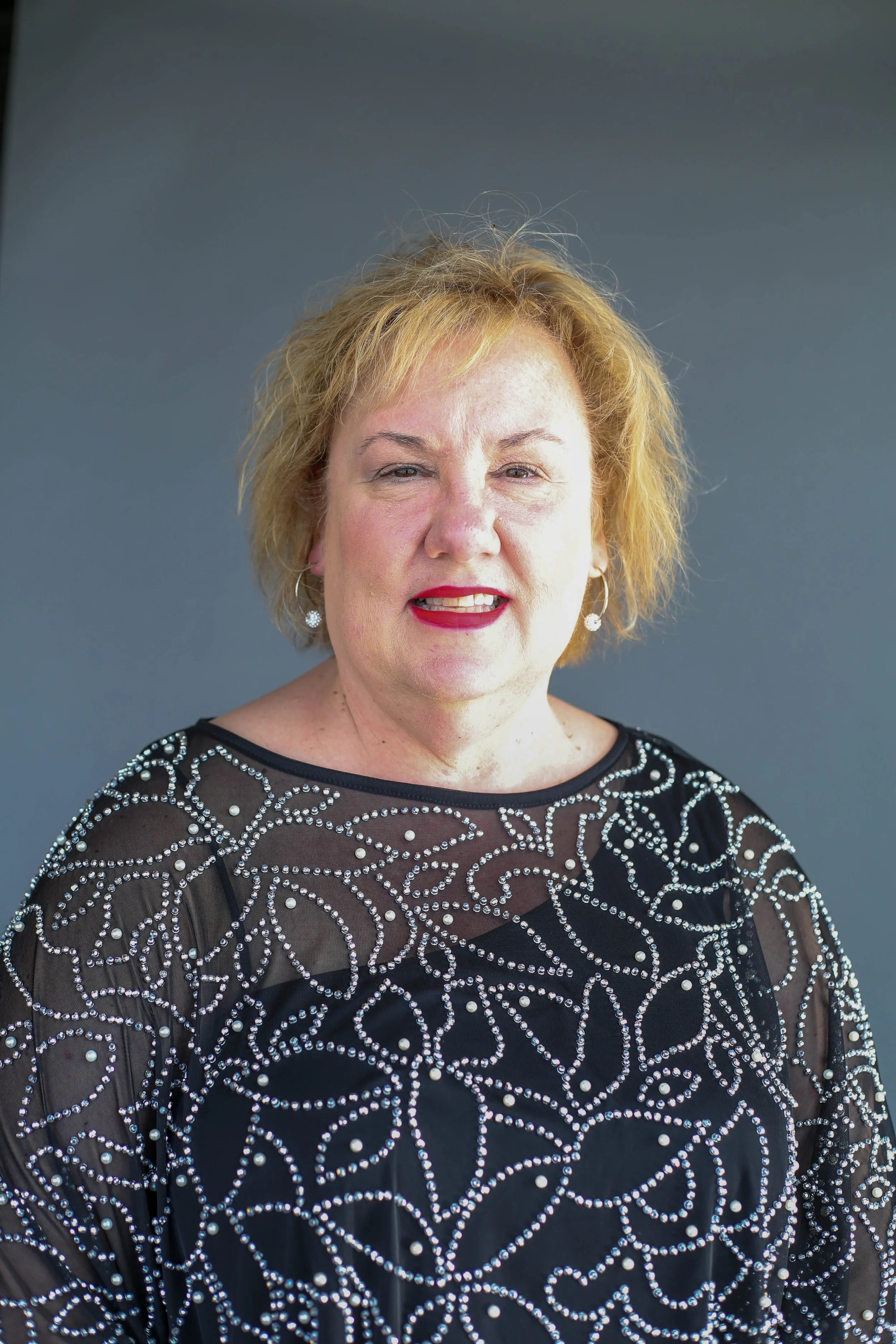 A woman with short blonde hair wearing a black top embroidered with silver beads and pearls, against a plain gray background.