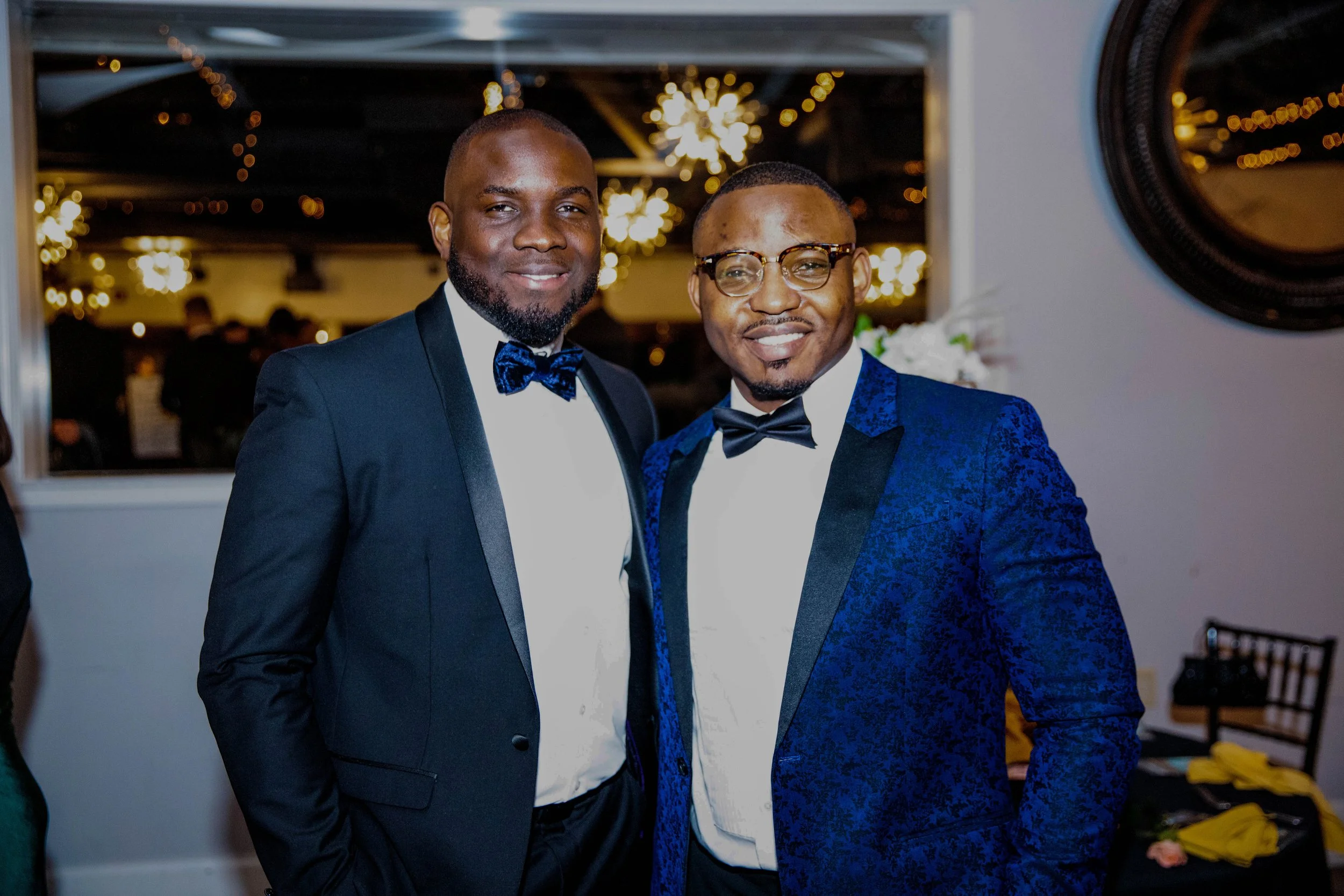 Two men in tuxedos smiling at a party or celebration, standing indoors with blurred background and decorative lighting.