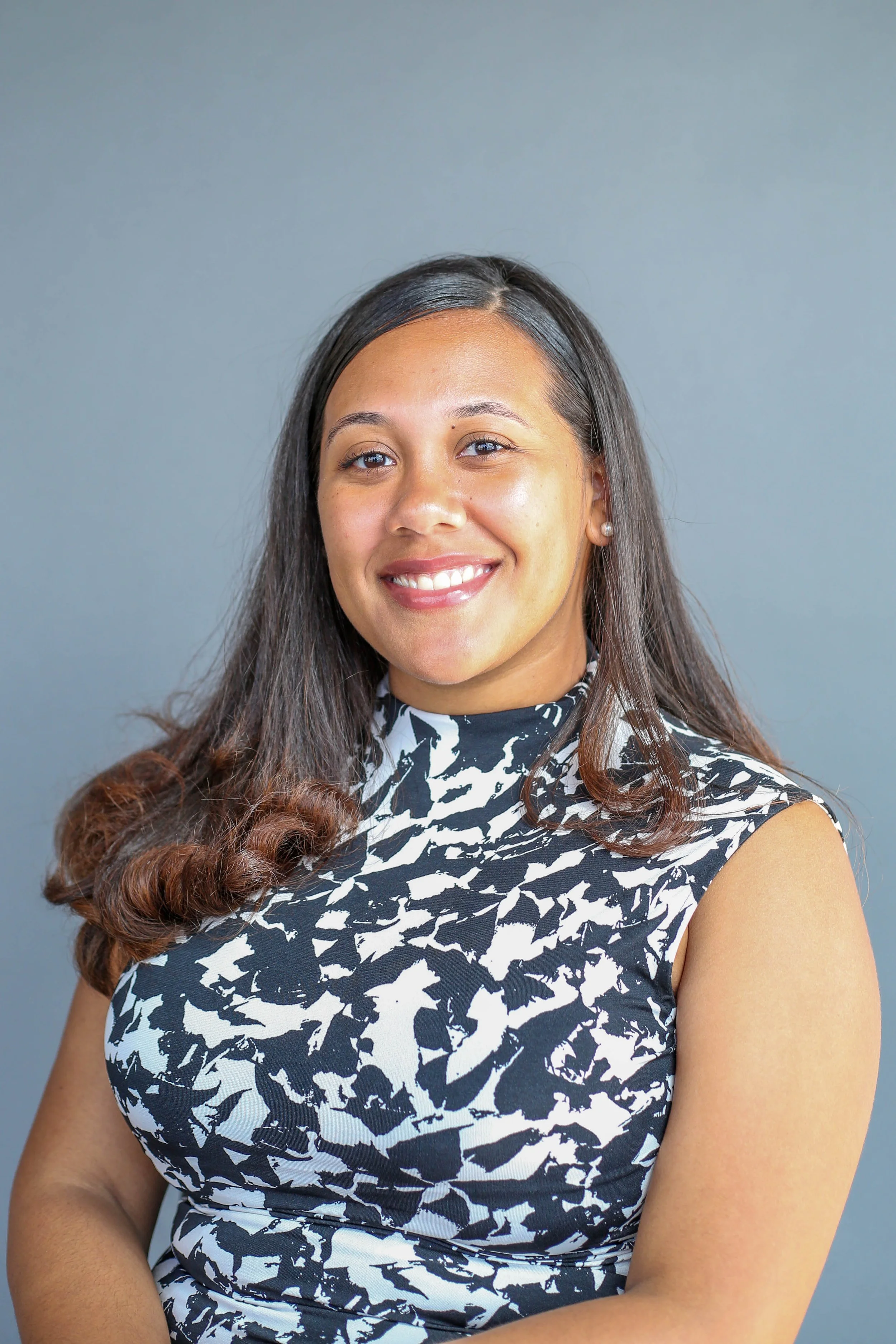 Close-up portrait of a young woman with long dark hair in a black and white patterned sleeveless dress, smiling against a plain gray background.