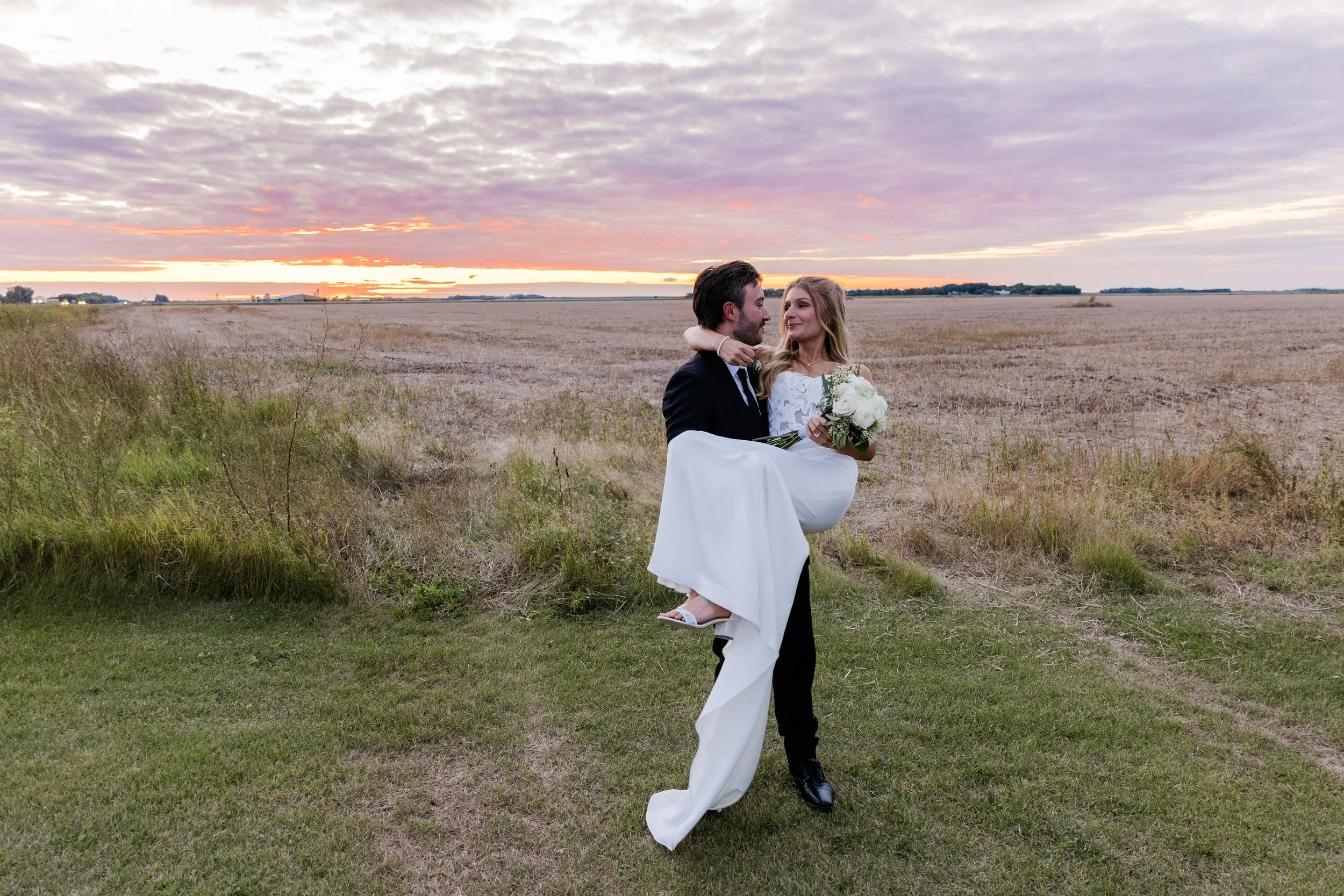 A groom carrying a bride in a white wedding dress across a grassy field during sunset, with a cloudy sky in the background.