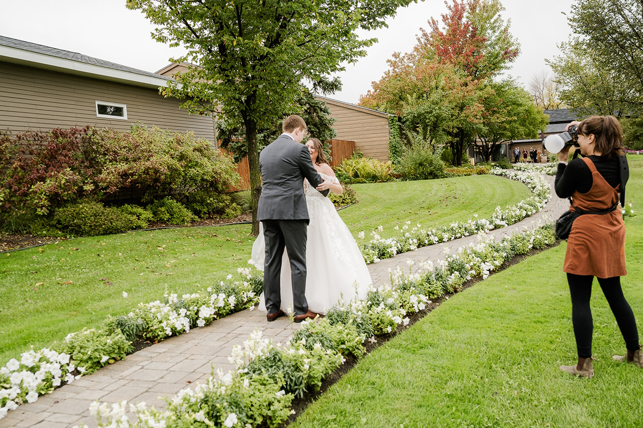 A bride and groom exchanging vows outdoors on a cloudy day, with a photographer capturing the moment on the right side of the image, surrounded by lush greenery and white flowers along the walkway.