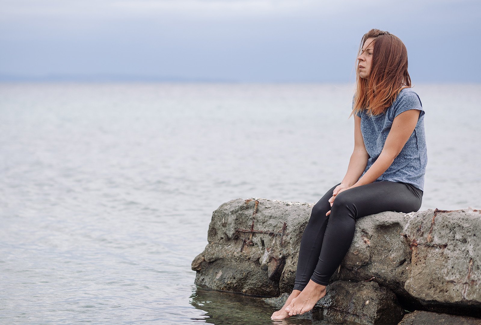 A young woman with auburn hair sitting on rocks at the edge of shore starring off toward the left with her face exposed.