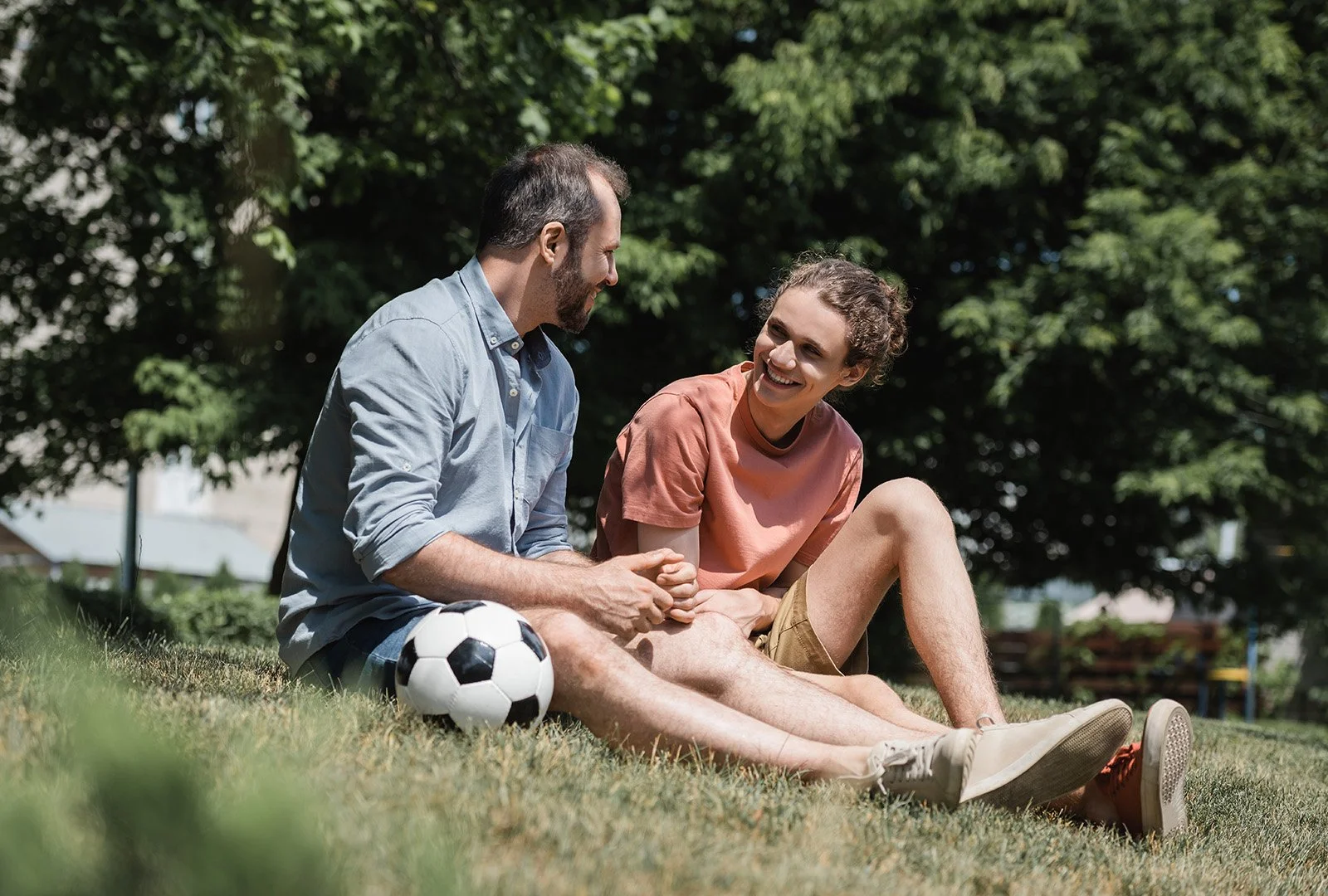 Dad and teenage son take a berak from Soccer to sit in the grass and talk.