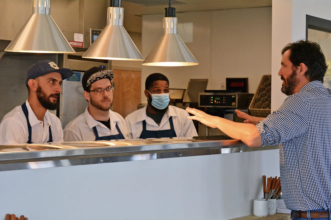 A sharp-dressed manager discusses the menu with kitchen crew in chef coats.