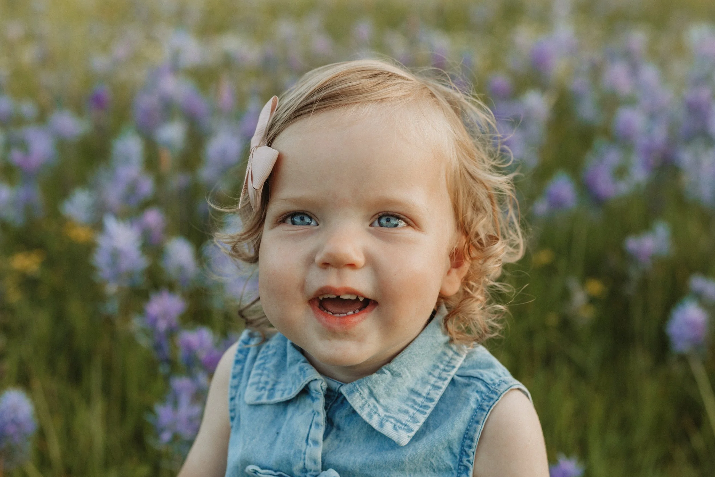 A young girl with blonde curly hair and blue eyes wearing a denim sleeveless top, smiling outdoors with purple flowers and green foliage in the background.