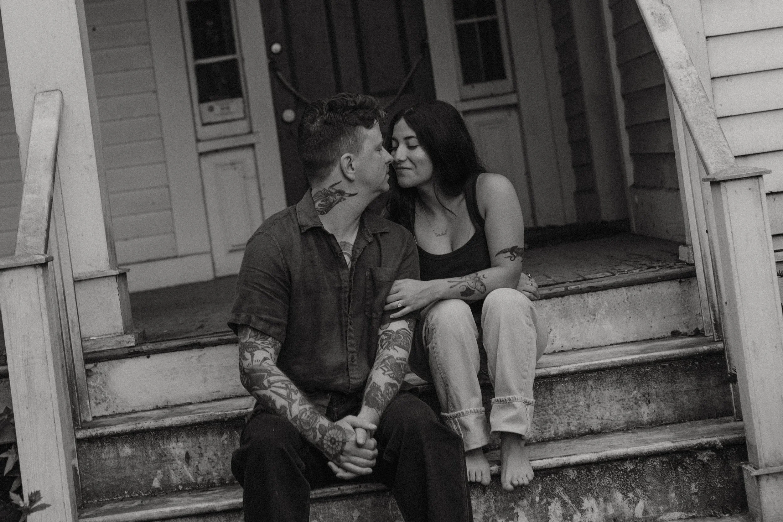 A black and white photo of a young couple sitting on the front steps of a house, leaning toward each other with foreheads touching, appearing intimate and affectionate.