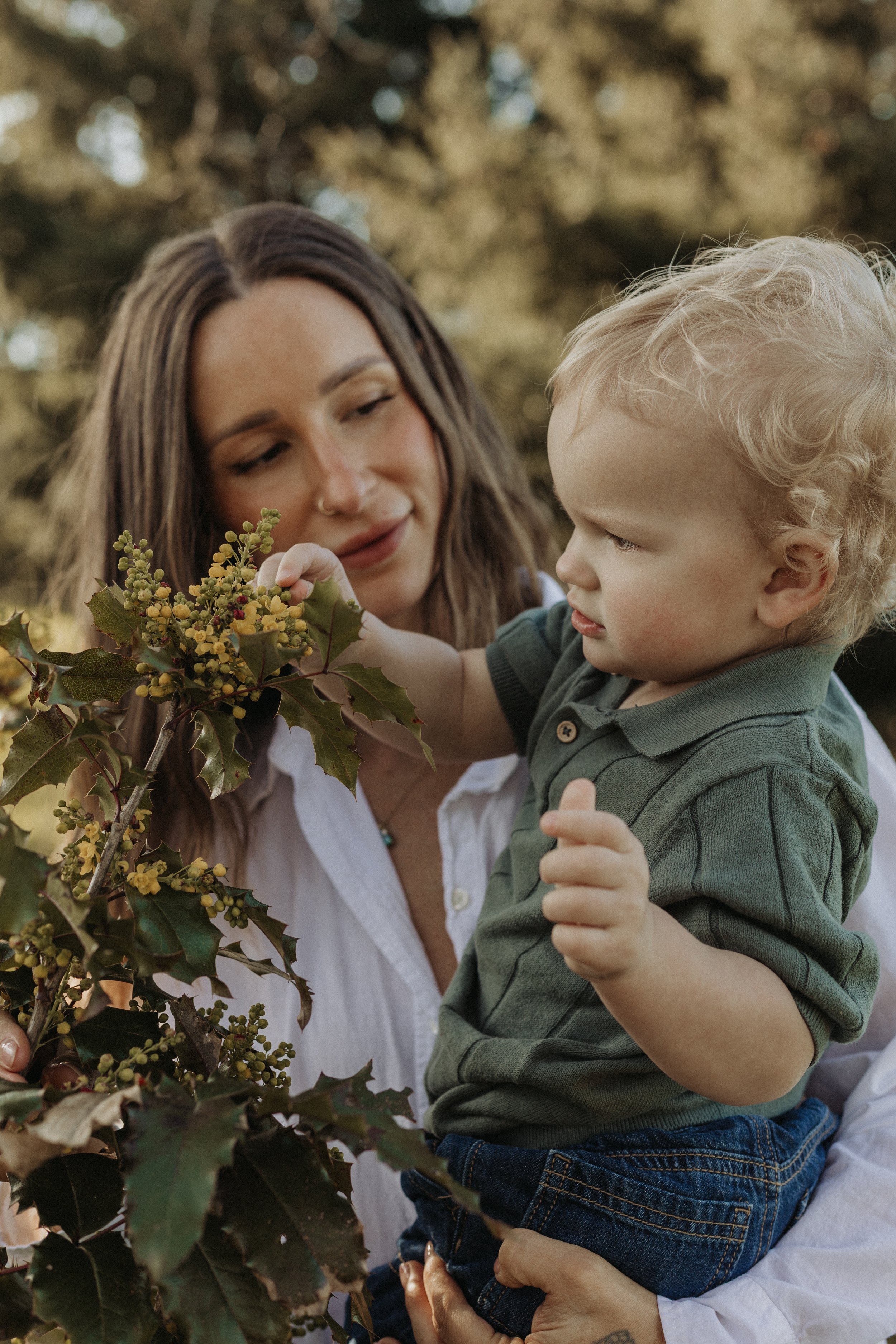 A woman with long brown hair and a nose ring holds a young blonde boy with curly hair, wearing a green collared shirt and jeans, as they look at a bunch of holly leaves and berries outdoors with a blurry background.