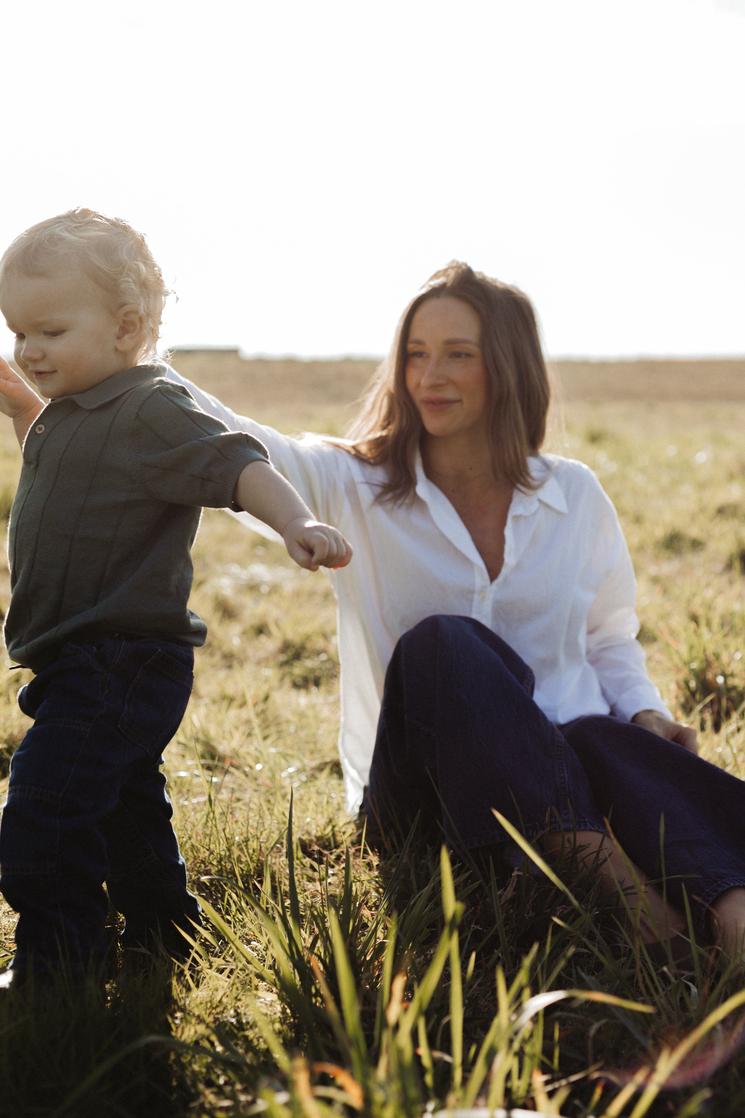 A woman with brown hair and a white shirt sitting on grass with a young blond boy standing in front of her, outdoors in a field during daytime.