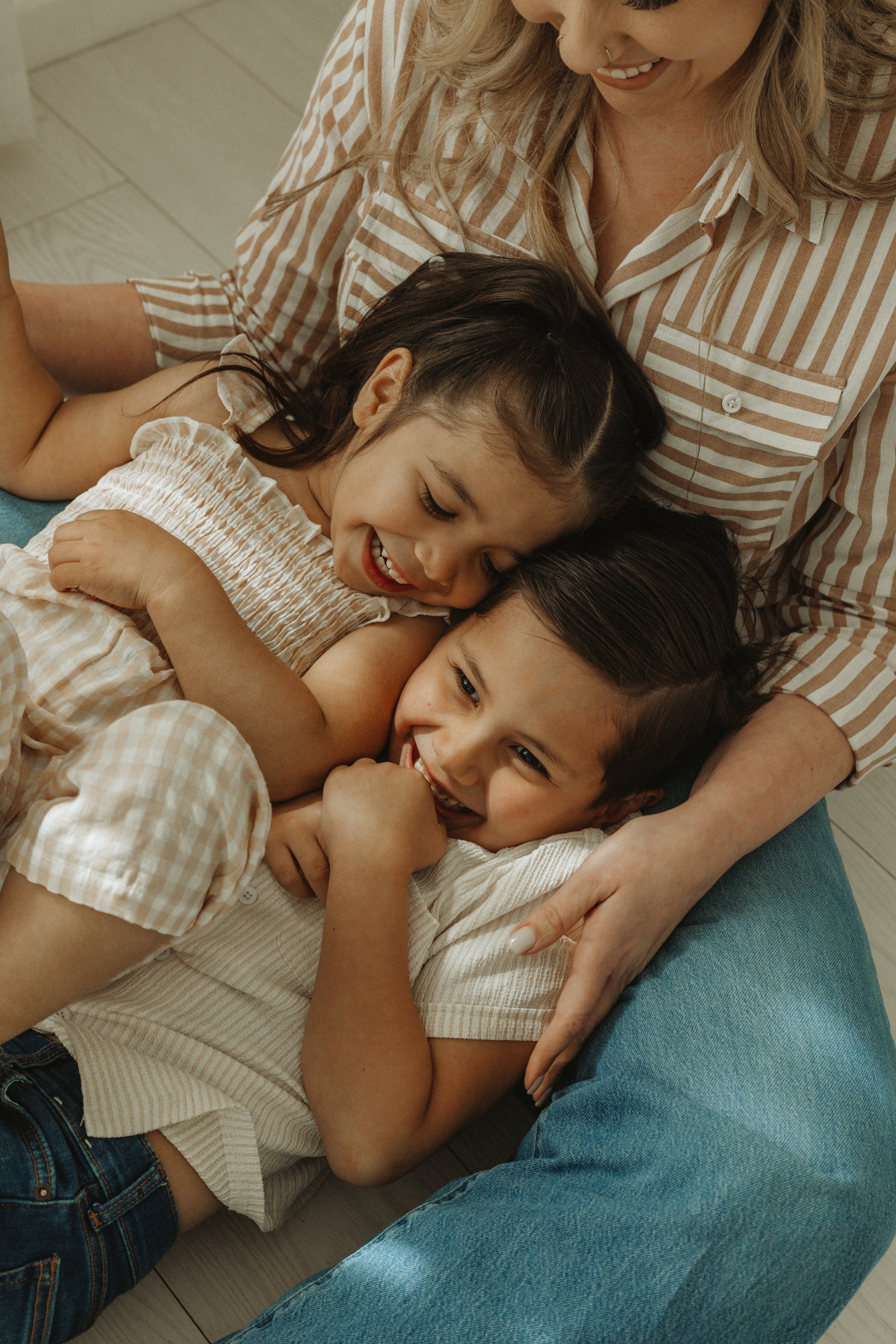 A woman with blonde hair and a septum piercing sits on the floor, smiling as she hugs two young girls who are laying on her lap. The girls are laughing and appear happy, sharing a moment of affection and joy.