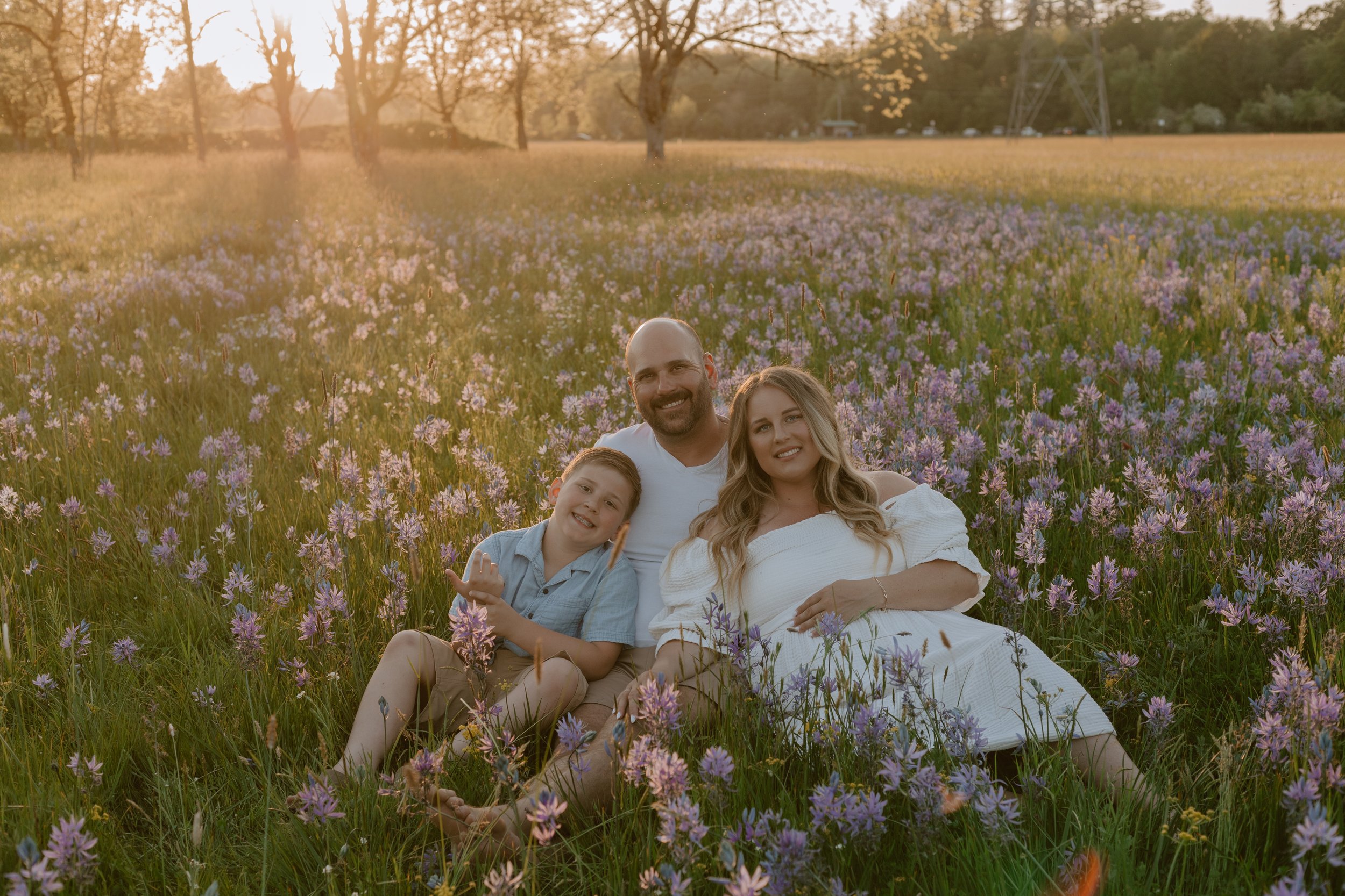 A family of four sitting and lying in a field of purple flowers during golden hour sunset with trees and power lines in the background.