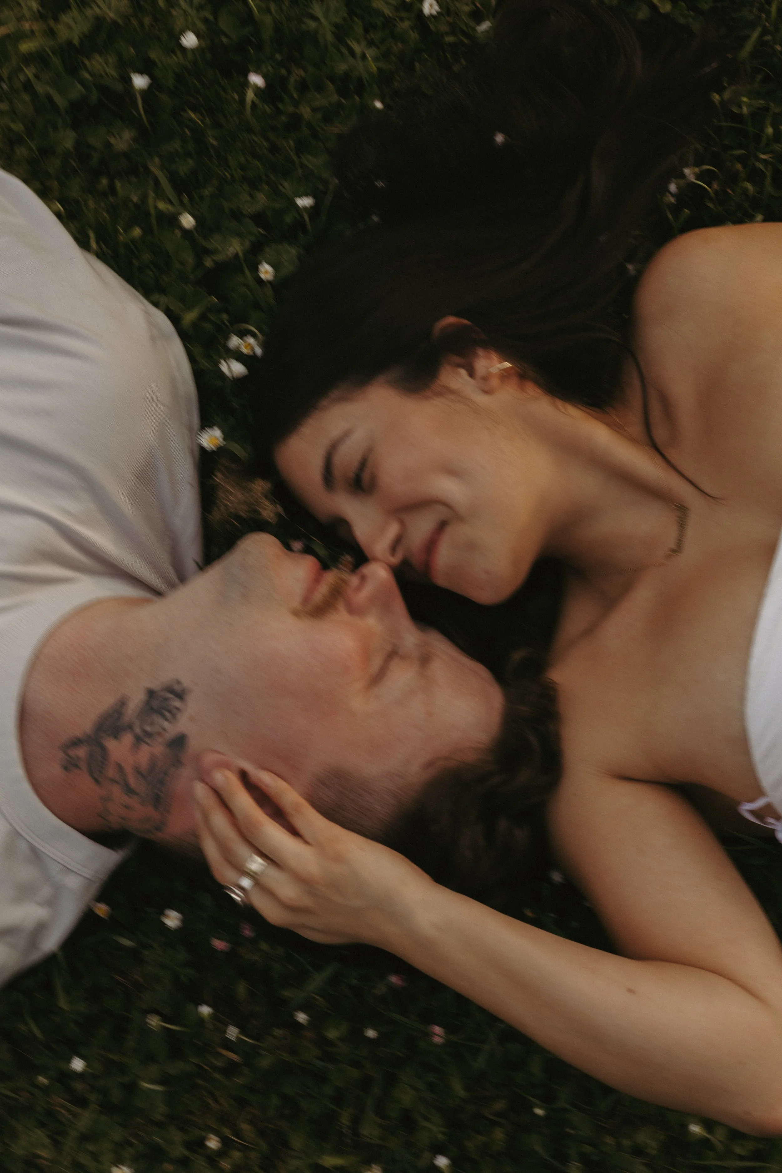 A couple lying on grass surrounded by small white flowers, the woman smiling with eyes closed, and the man with a tattoo on his head, holding her hand gently.