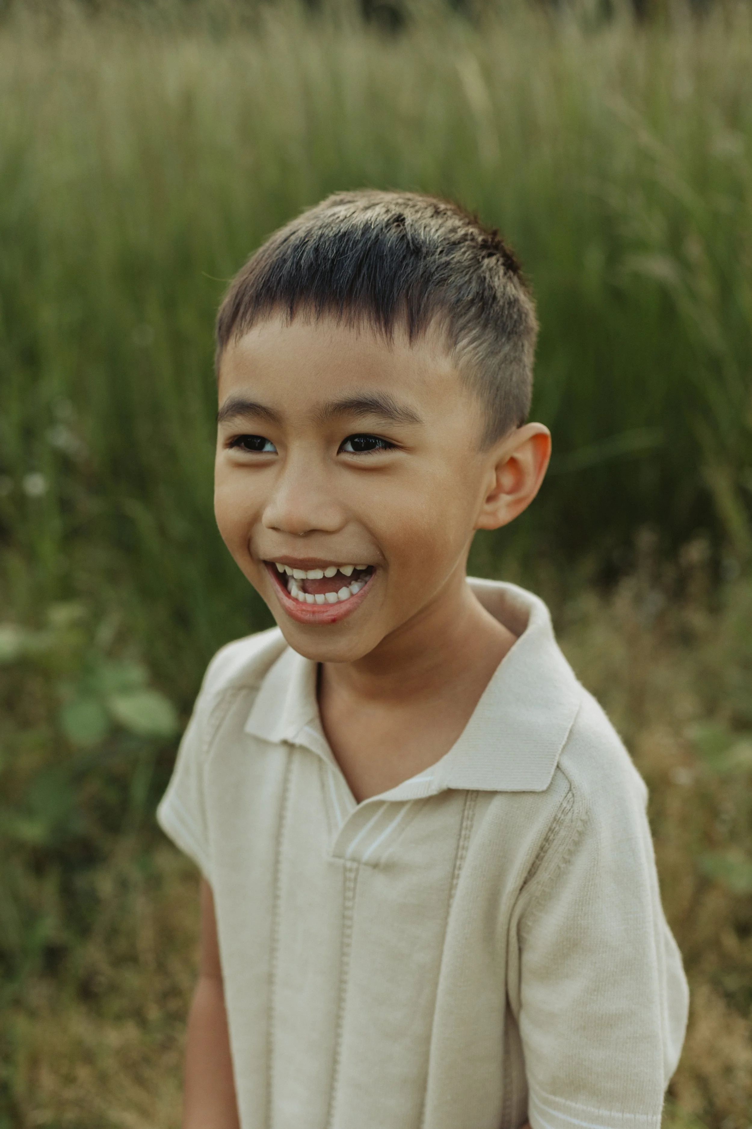 Close-up of a young boy with short dark hair smiling outdoors with greenery in the background.