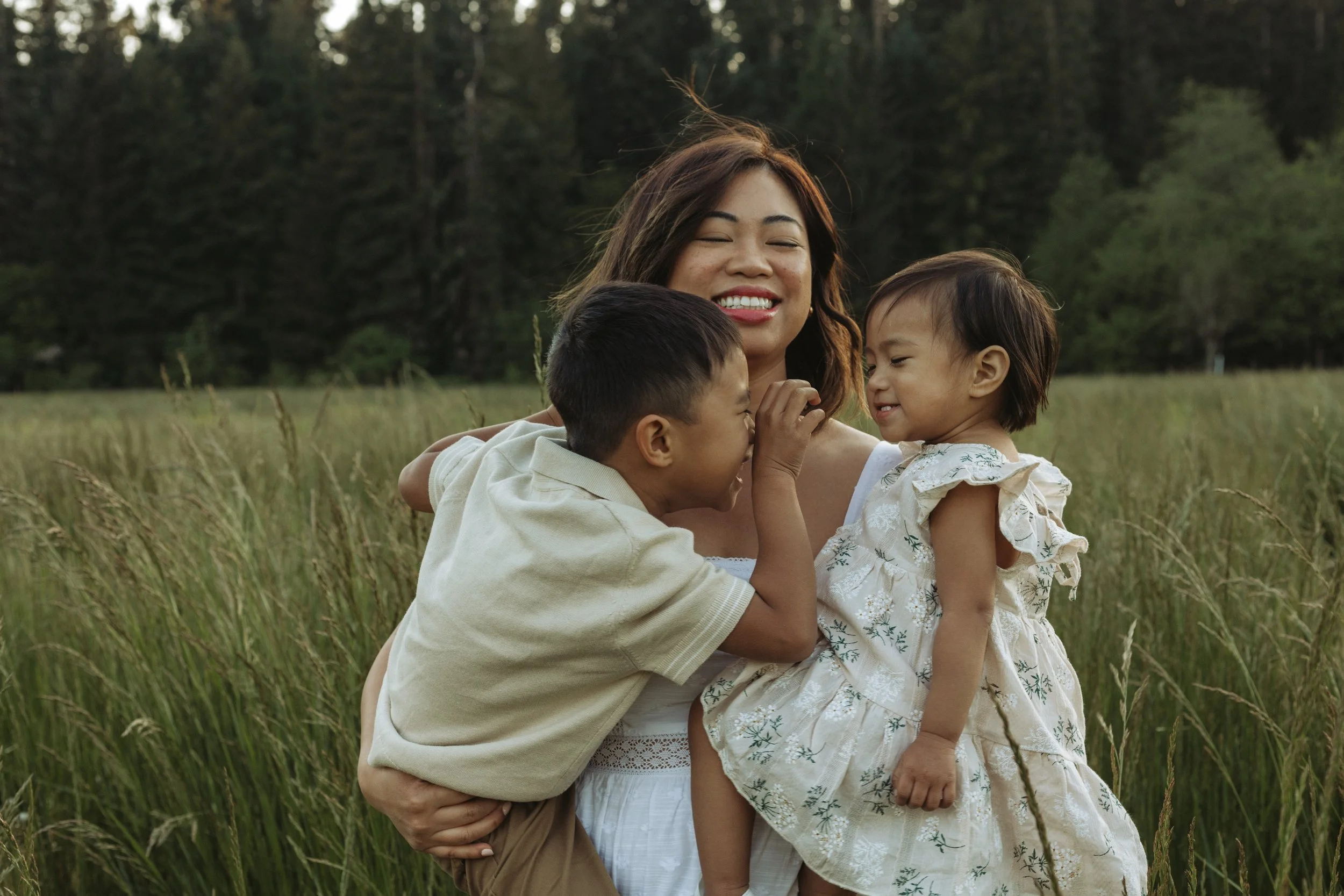 A woman with long brown hair smiling and holding two young children in a grassy field surrounded by trees.