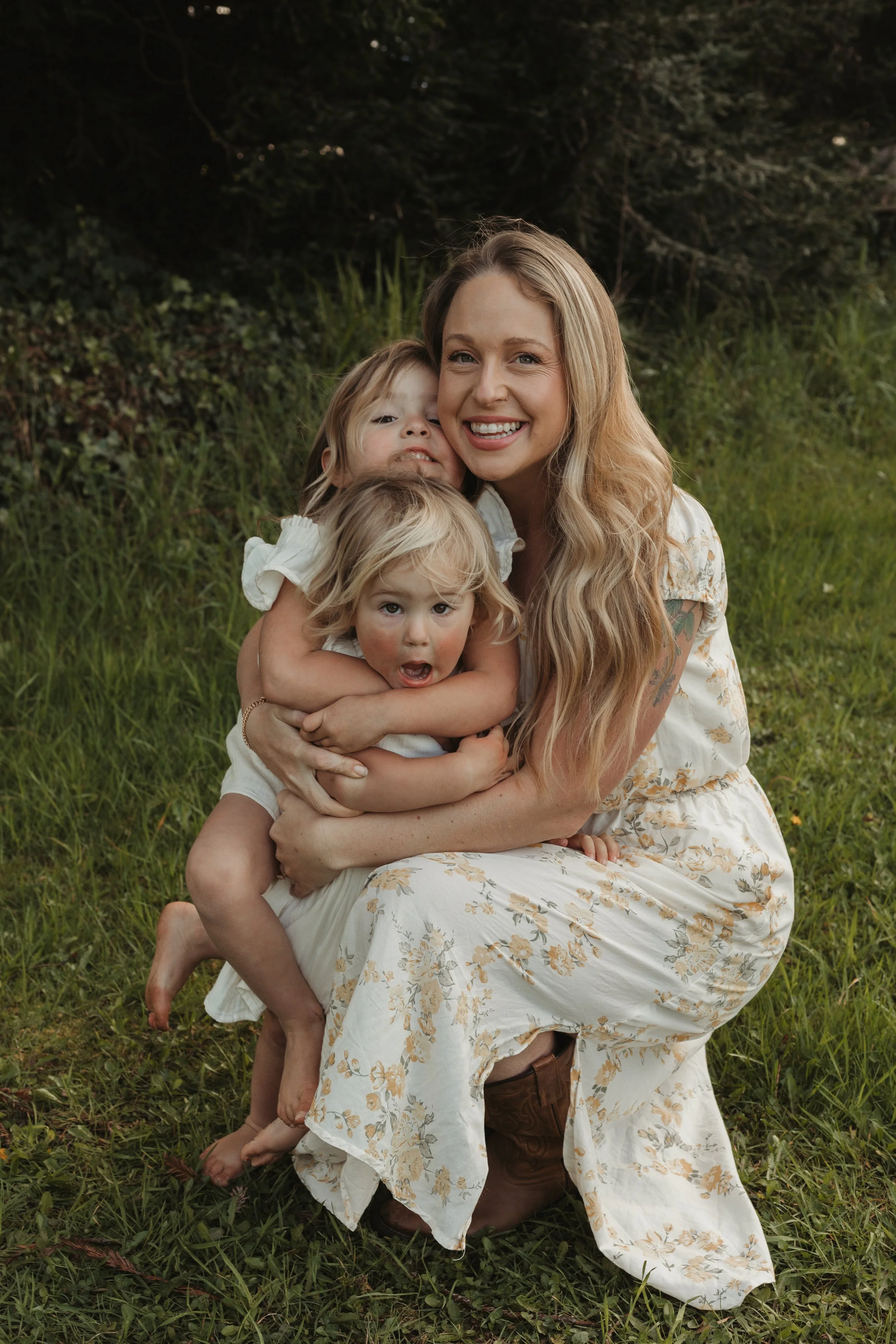 A woman with long blonde hair sitting on the grass outdoors, hugging three young children who are leaning on her, with a background of trees and greenery.