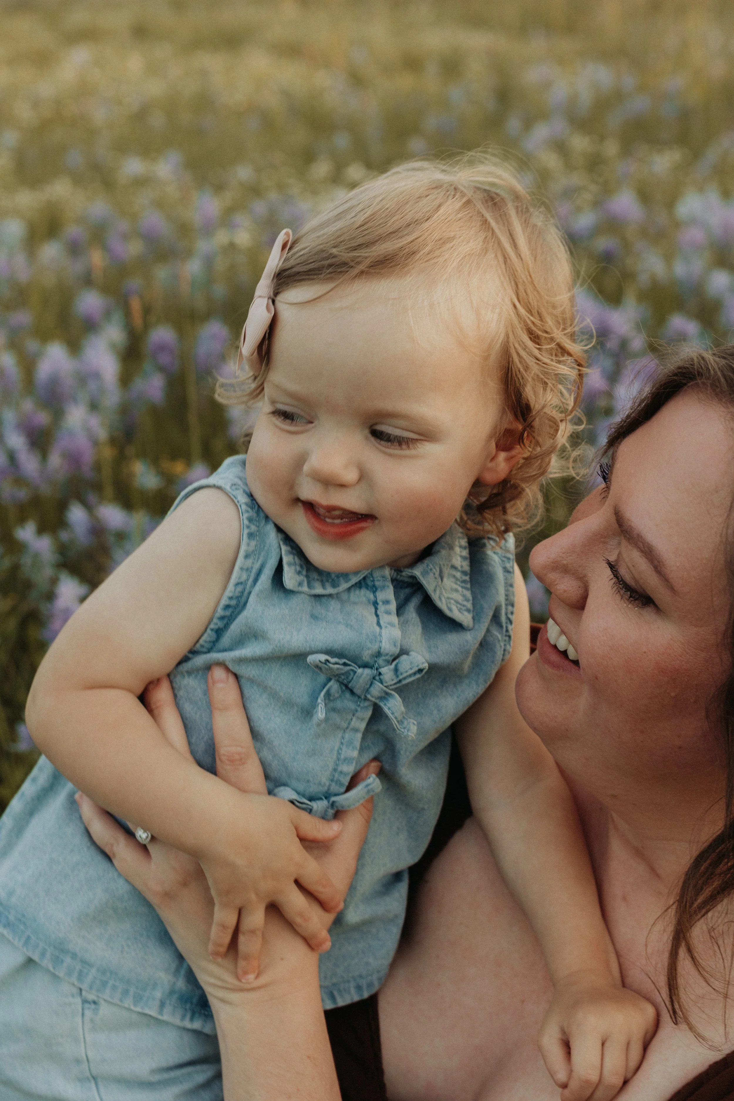 A woman holding a smiling young girl with curly blonde hair, wearing a denim dress, outdoors with purple flowers in the background.