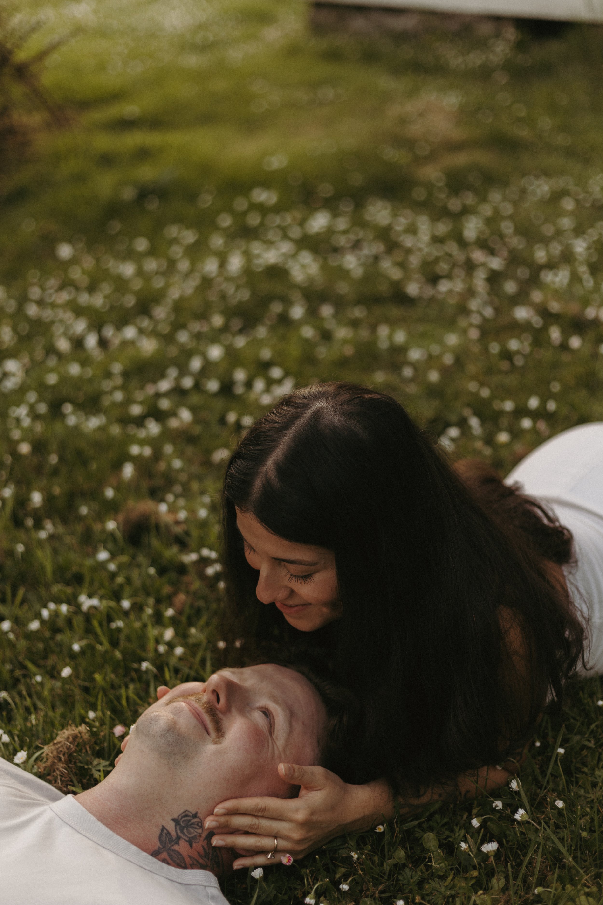 A woman with long dark hair leaning over and kissing a man with a shaved head lying on the grass, surrounded by small white flowers.