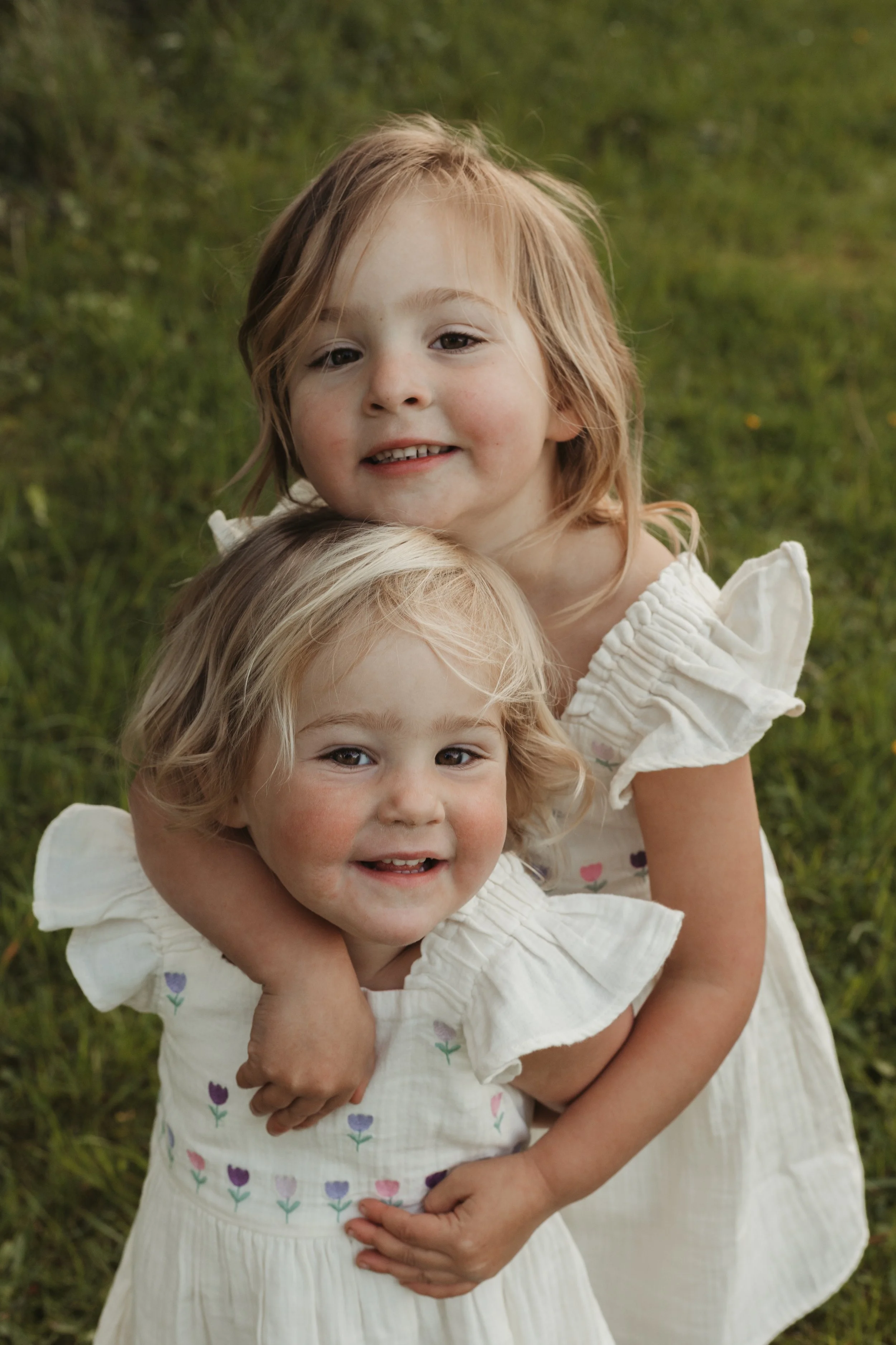Two young girls with light brown hair, wearing white dresses with colorful flower patterns, embracing each other outdoors on grass.