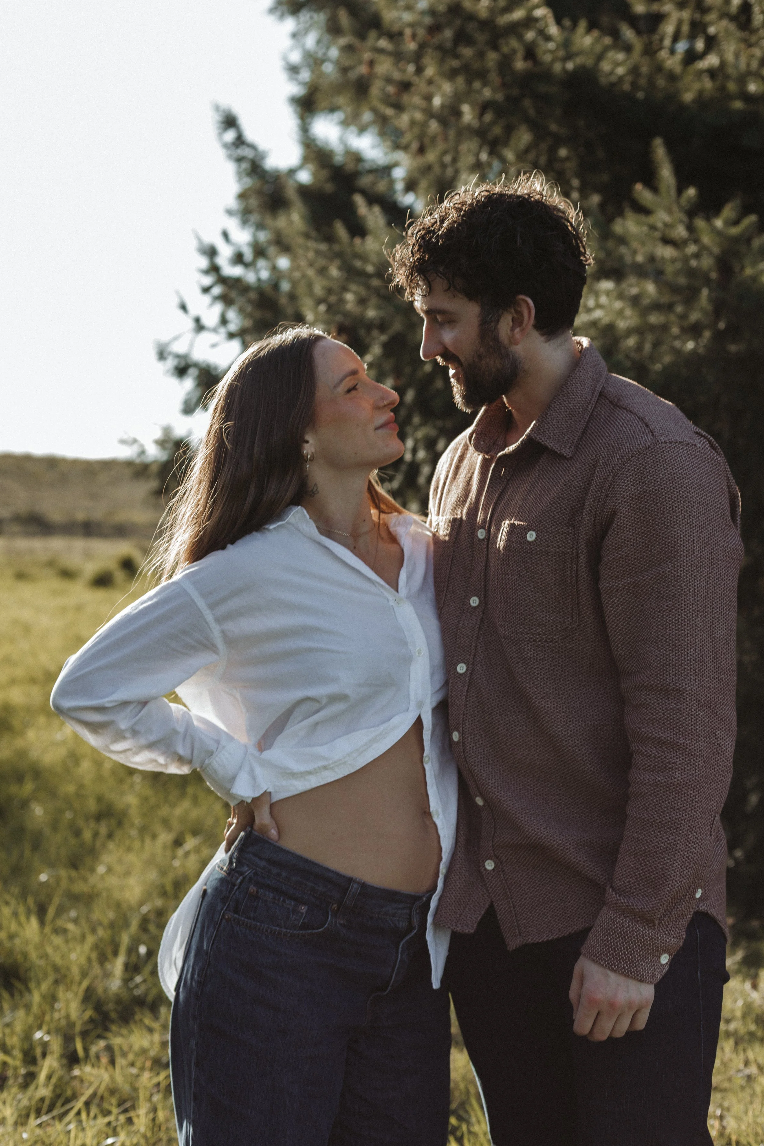 A man and woman stand close together outdoors, smiling and looking at each other. The woman wears a white shirt, unbuttoned at the bottom, and jeans. The man wears a brown shirt. They stand in a grassy field with trees in the background during the da
