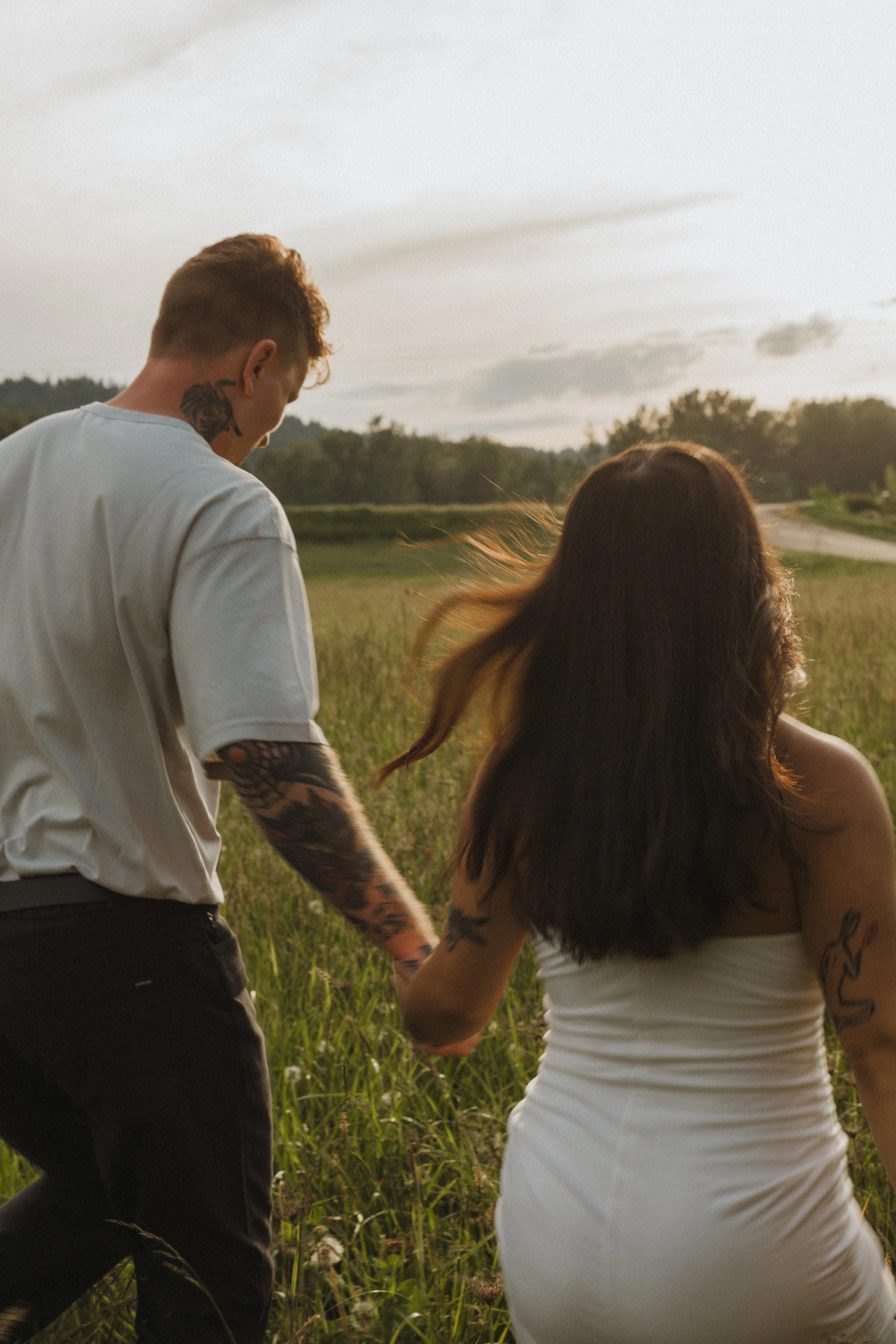 A young man with tattoos on his neck and arms, and a woman with long dark hair and tattoos on her arms, are standing in a grassy field during sunset. They are holding hands and facing away from the camera.