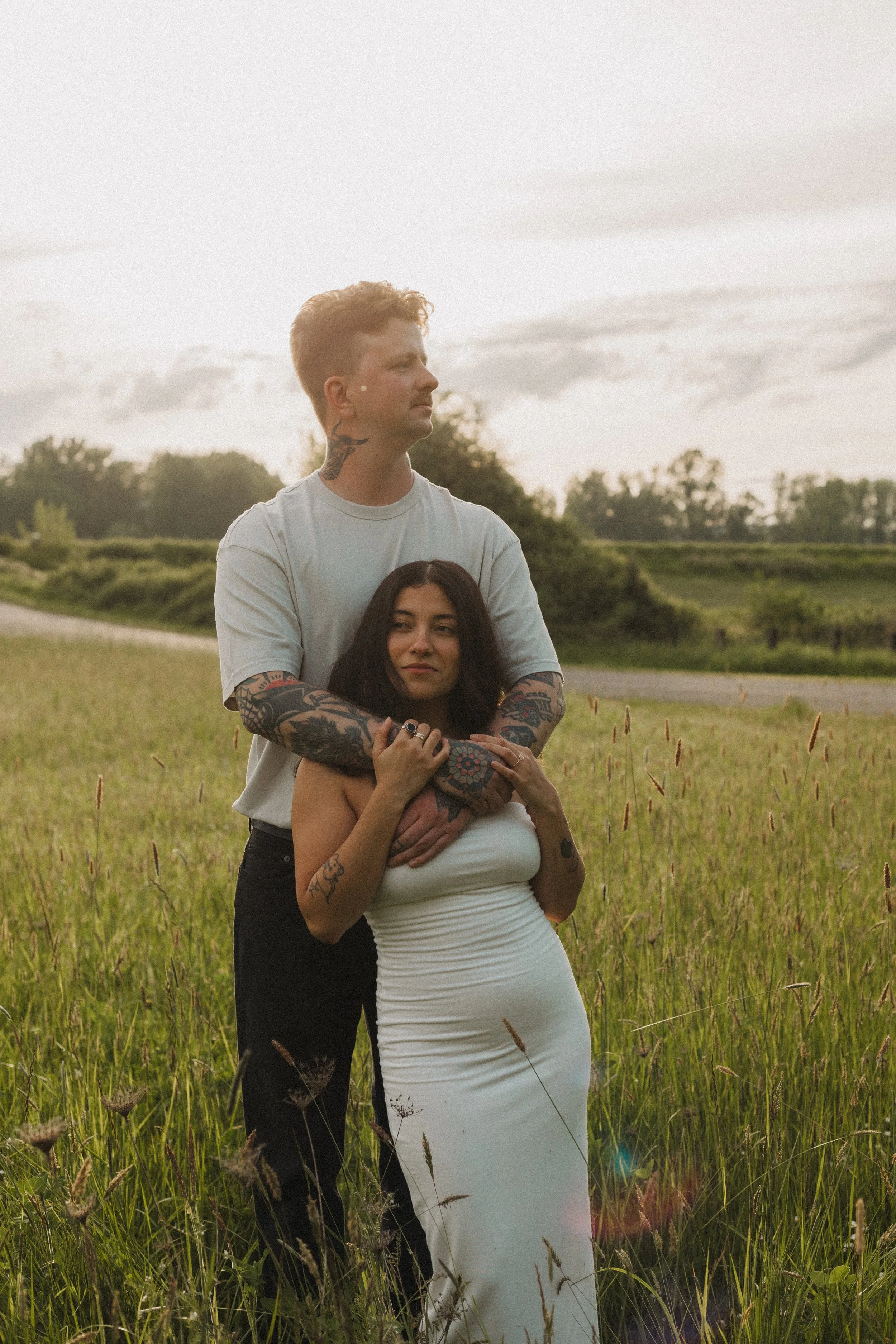 A man with tattoos and a woman in a white dress standing in a grassy field during sunset, with trees and a cloudy sky in the background.