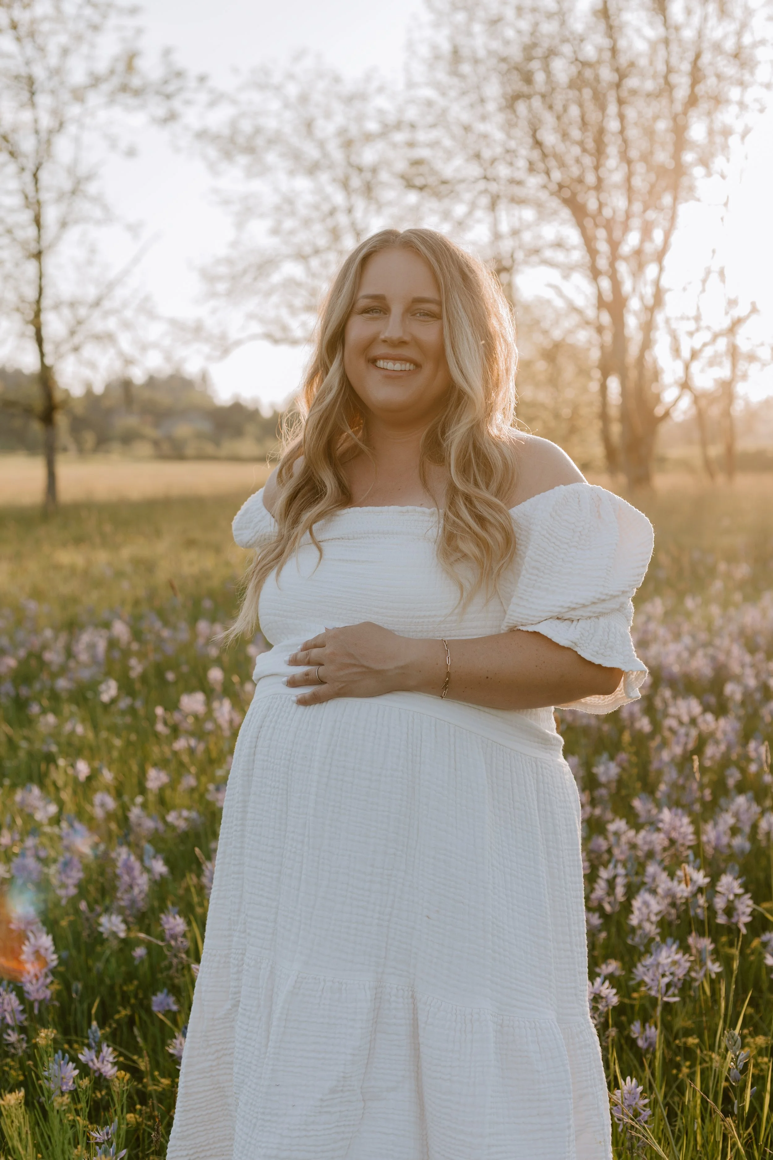 A woman wearing a white dress smiling in a field of purple flowers with trees and sunlight in the background.