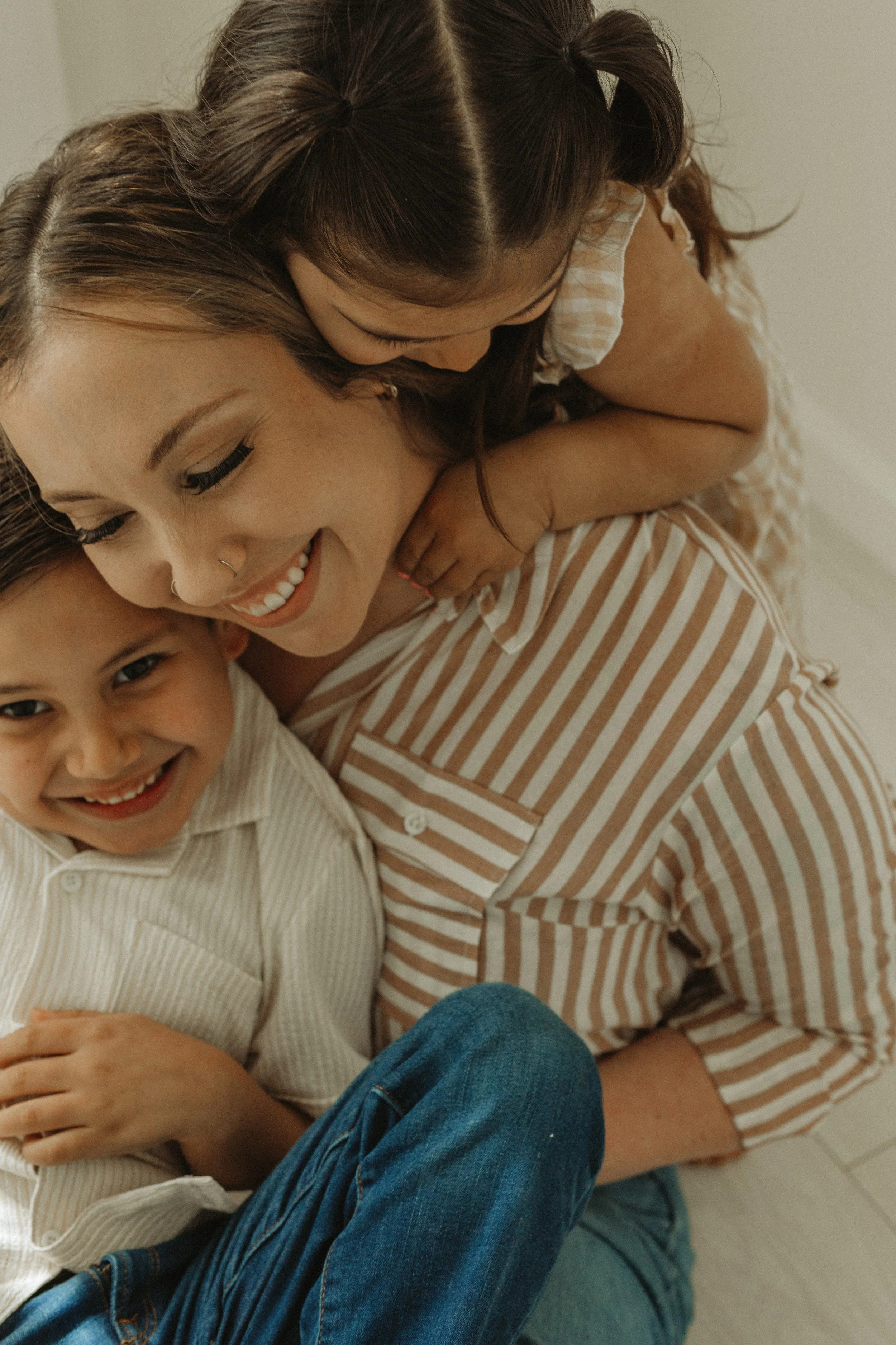 A woman with long hair leaning over and hugging a child with a big smile. The woman is wearing a striped shirt and the child is wearing a light-colored button-up shirt. The scene is warm and loving, in a cozy indoor setting.