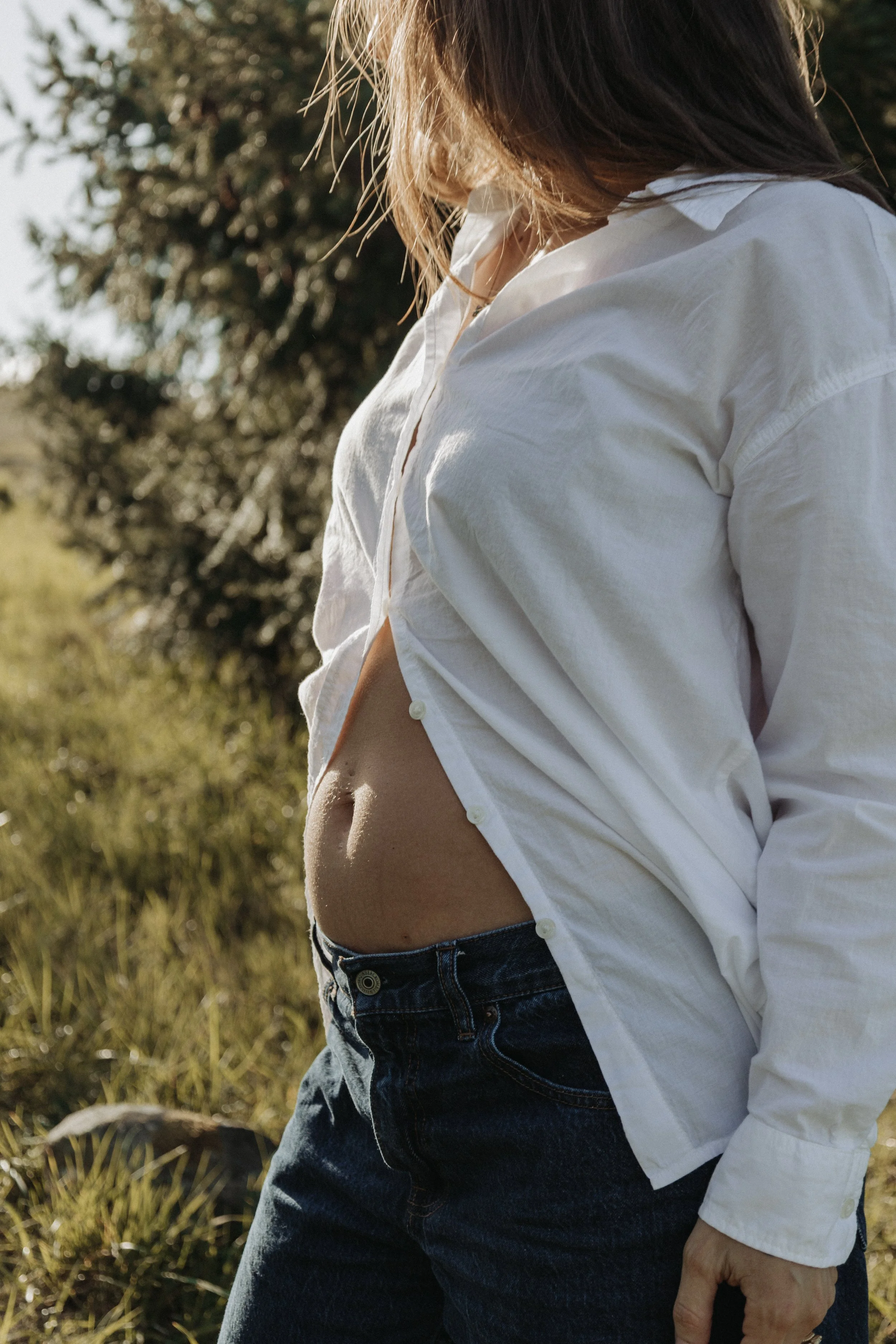 Close-up of a person wearing a white shirt and jeans, standing outdoors in a grassy area with trees in the background, with sunlight illuminating the scene.
