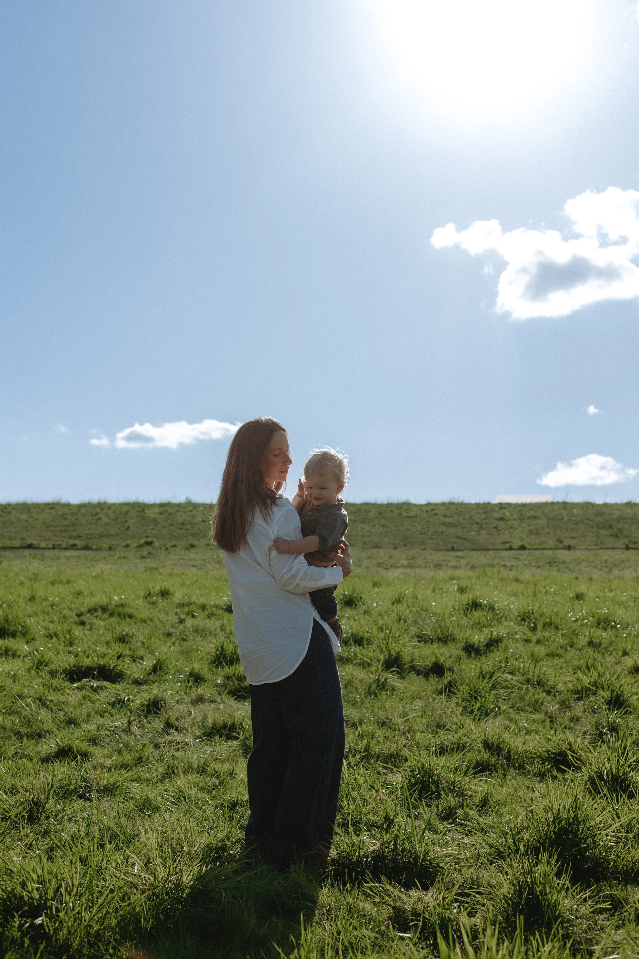 A woman holding a young child in an open grassy field under a bright blue sky with scattered clouds and the sun shining.