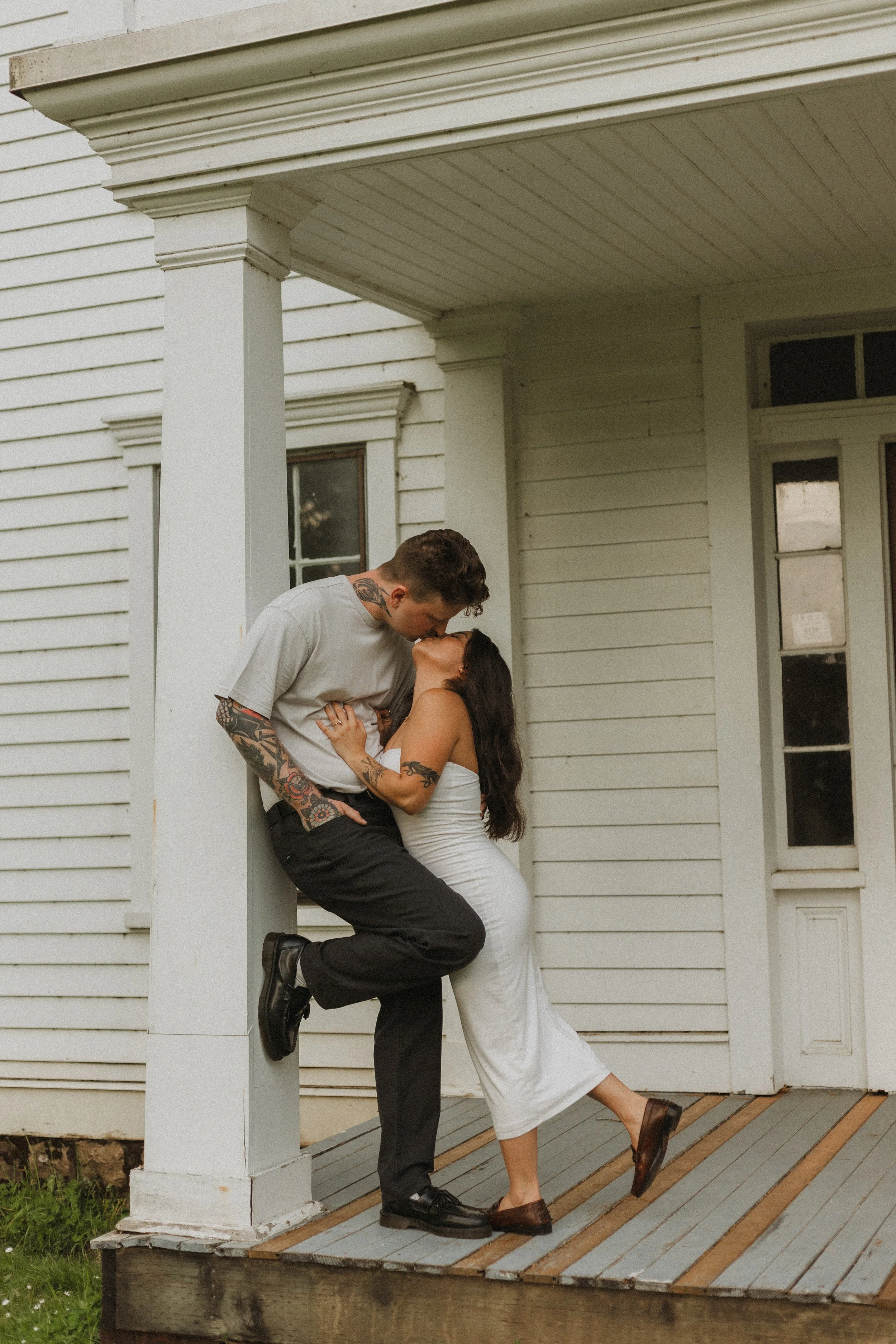 A couple shares a romantic moment on the porch of a white wooden house. The man is dressed in a casual gray t-shirt and black pants, leaning against the porch column. The woman, dressed in a white dress, stands on her tiptoes, leaning into the man's 