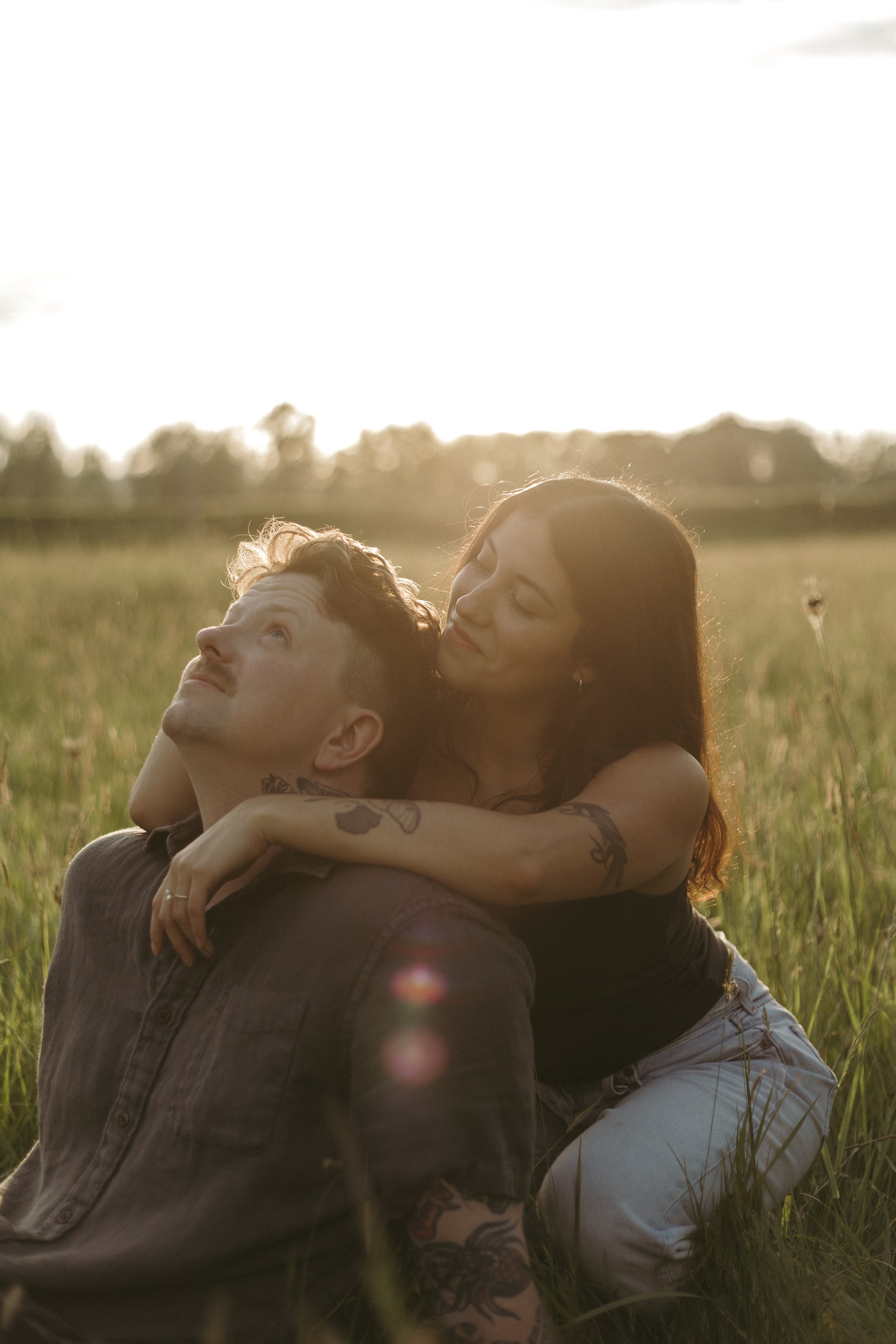 A couple sitting in a grassy field during sunset, with the woman hugging the man from behind, both smiling and enjoying each other's company.