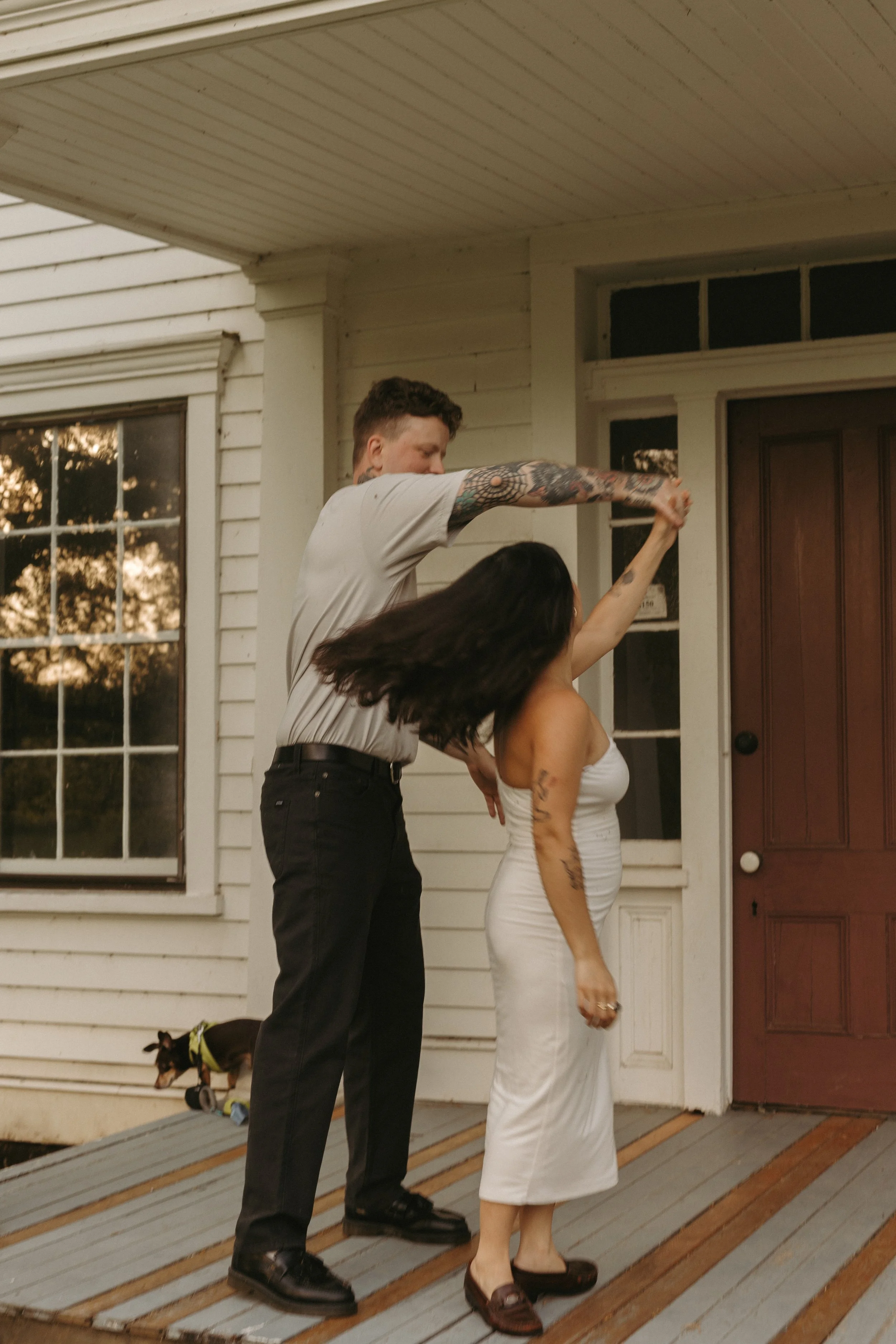 A man with tattoos on his arms and short hair appears to be throwing punch at a woman's face with black hair. The woman, dressed in a white dress, seems to be a mannequin. The scene is set outside a house with a door and large windows. There is a sma