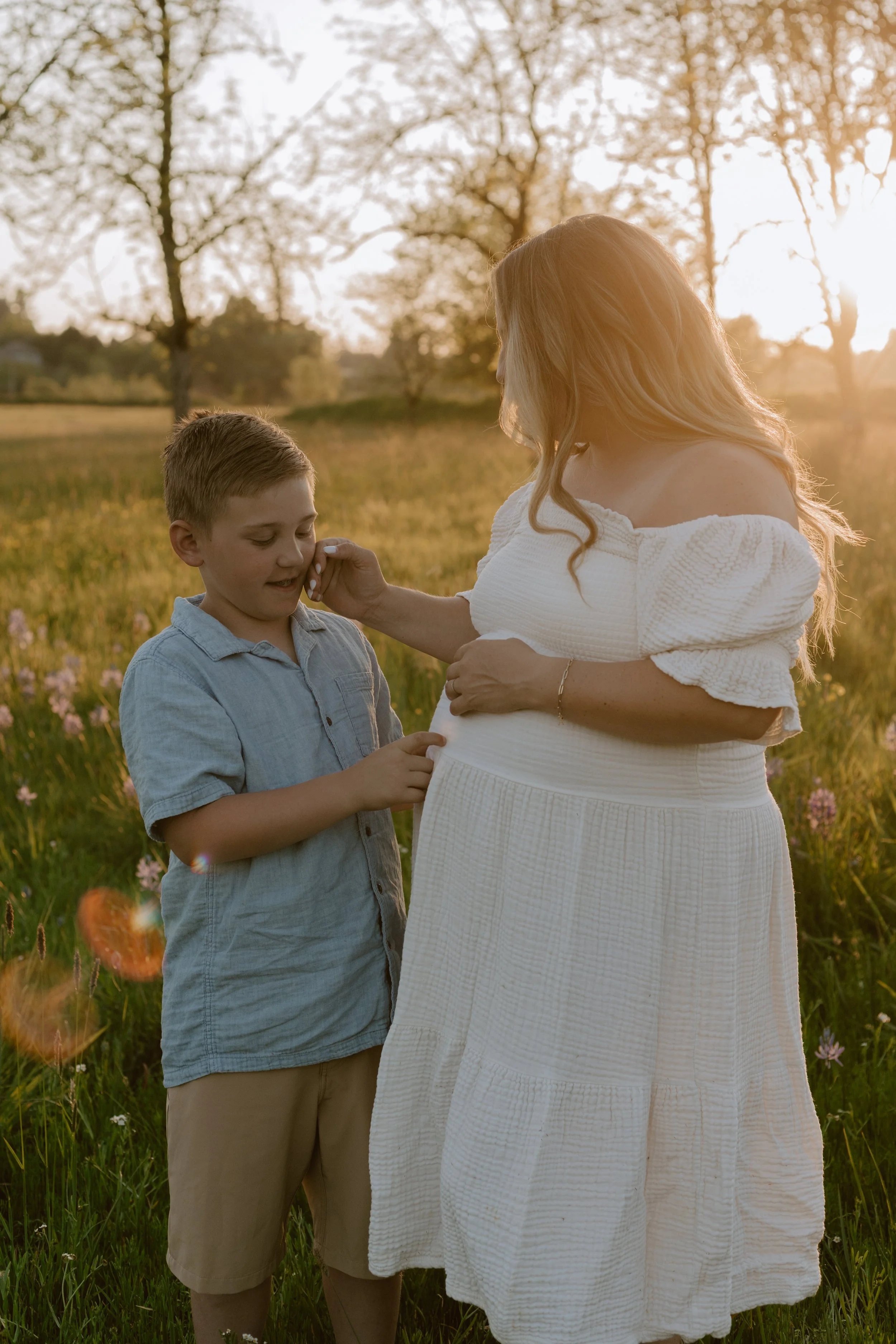 A pregnant woman with long, loose hair in a white dress standing in a field with a young boy during sunset, with trees and flowers in the background.