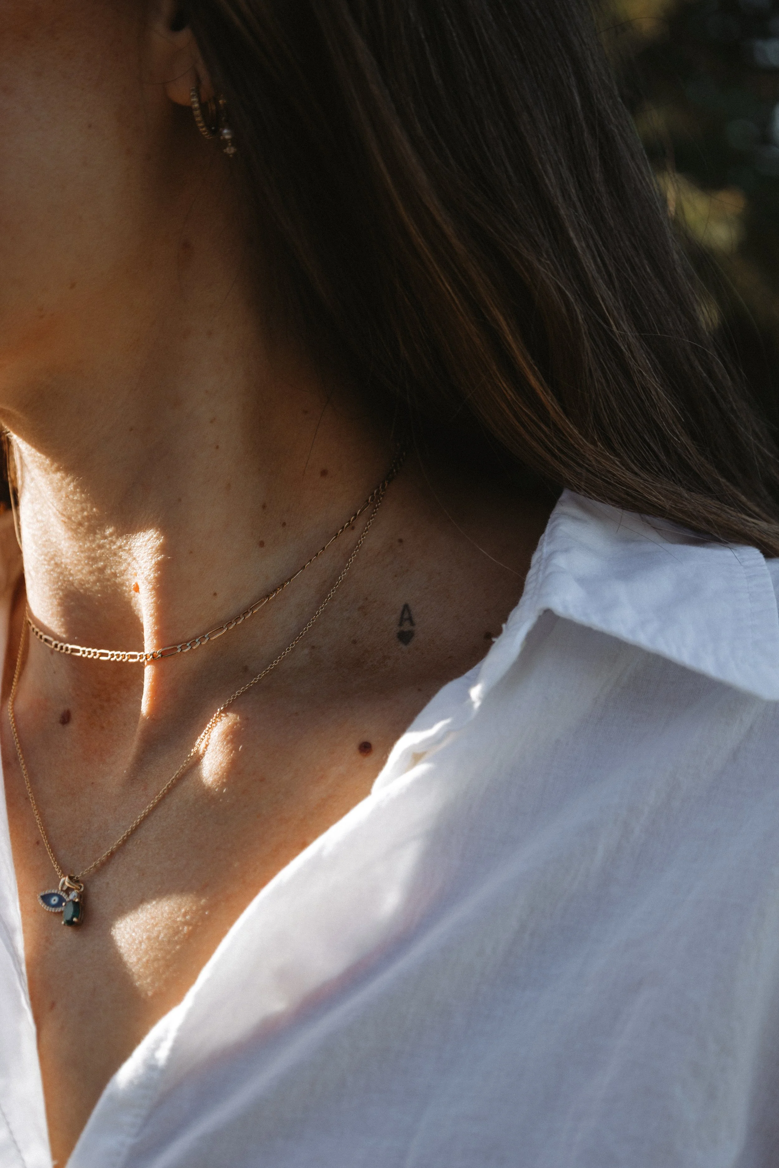 Close-up of a woman's neck and shoulder with layered gold necklaces, a small tattoo of a heart and the letter A, and earrings; she is wearing a white shirt.