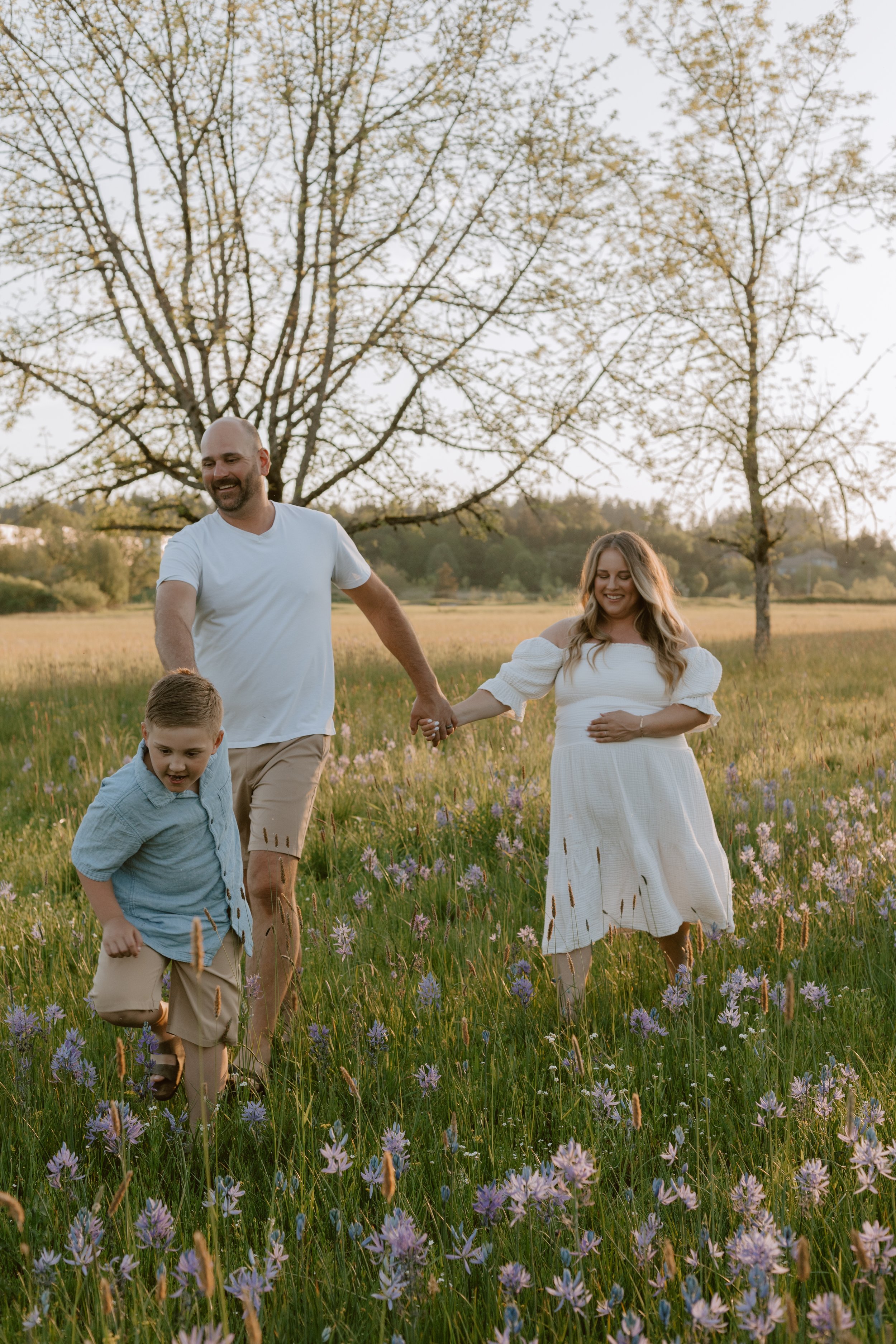 A family of four, including a man, woman, and two children, walks hand in hand through a field of purple wildflowers during sunset or late afternoon, with trees and a clear sky in the background.