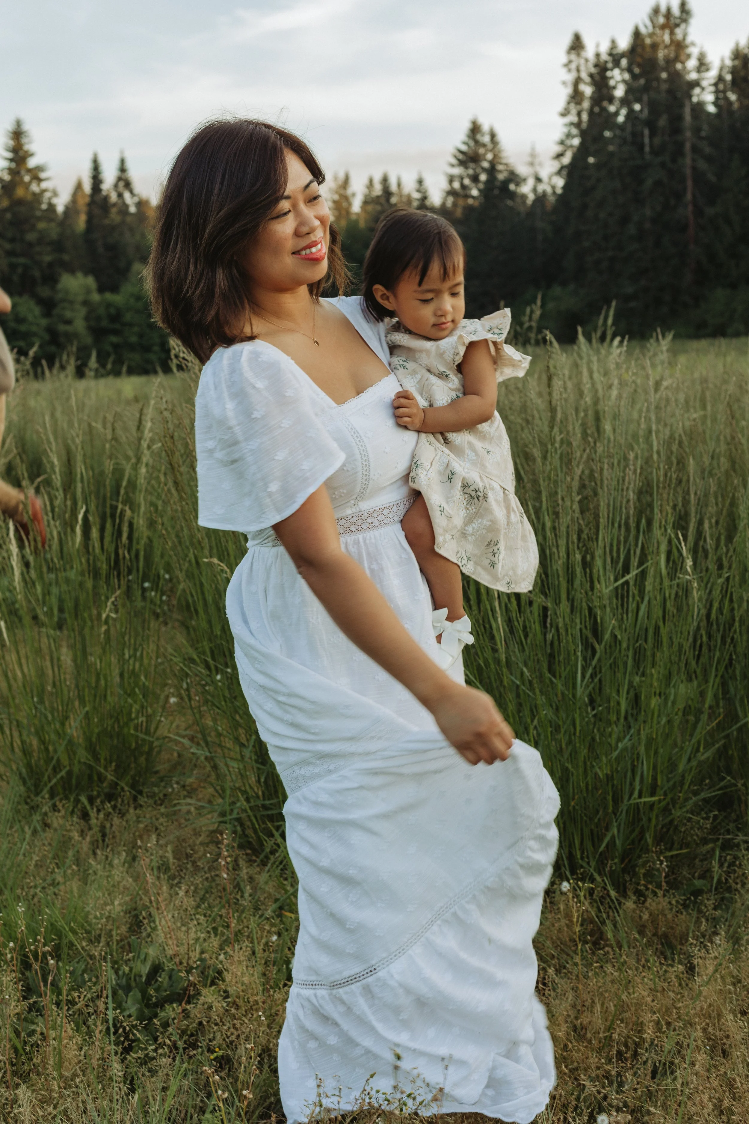 A woman holding a young girl in a grassy field surrounded by trees during daylight. The woman is smiling, wearing a white dress, and has shoulder-length dark hair. The girl is wearing a floral dress and has dark hair. Both appear to be enjoying a ser
