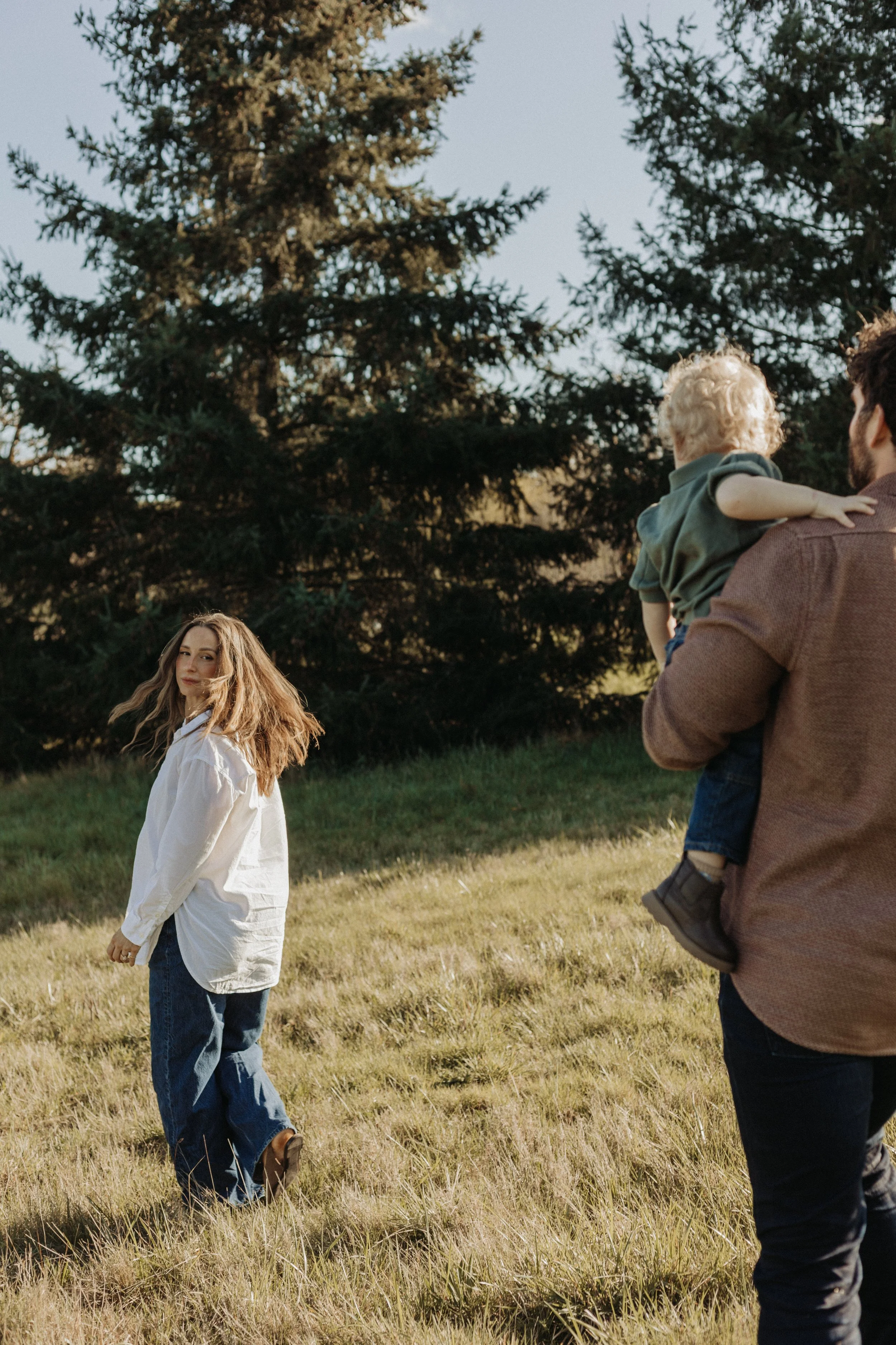 A man holding a young child and a woman walking through a grassy field with large trees in the background during daylight.