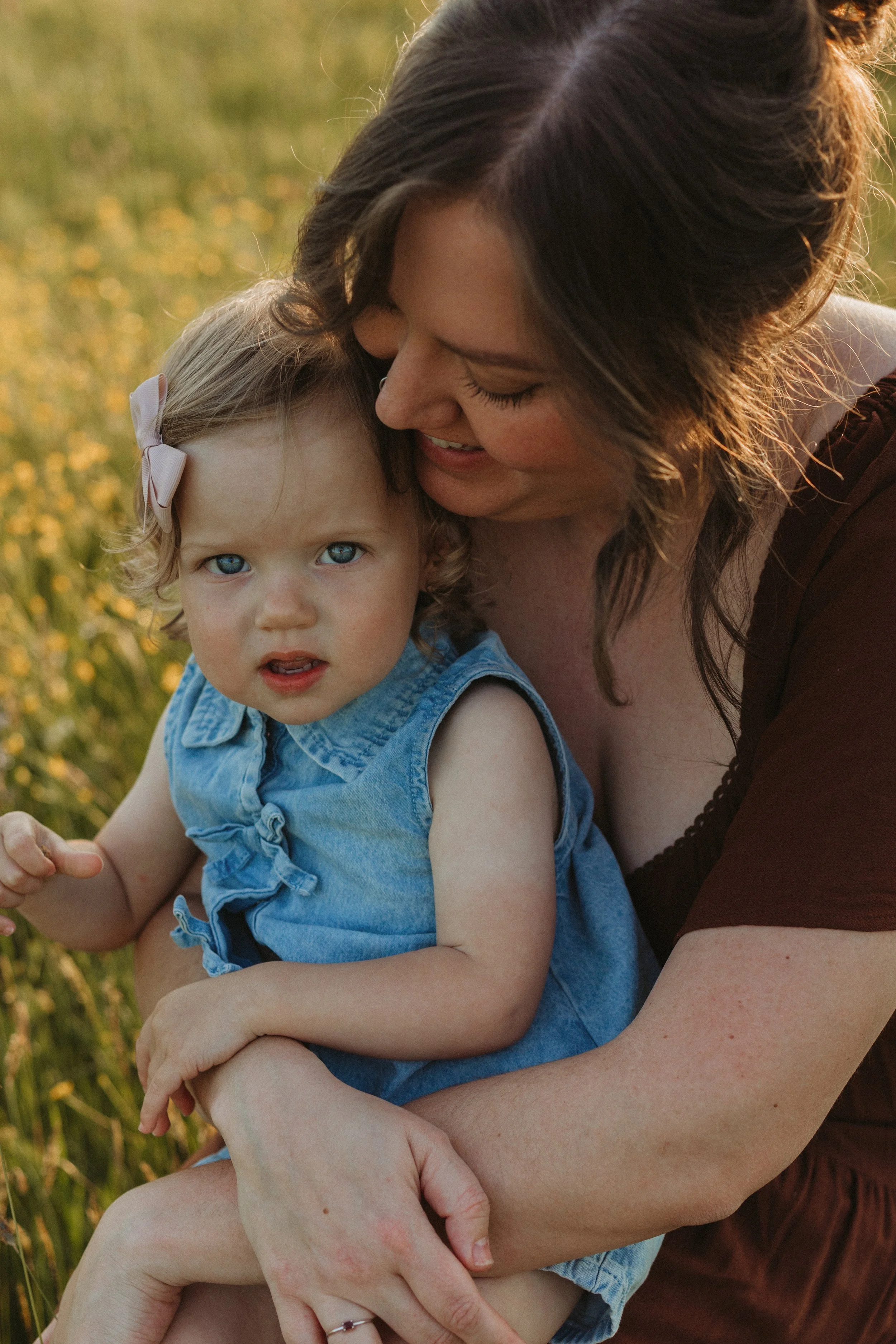 A woman with brown hair smiling and embracing a young girl with blonde hair and blue eyes in a field of yellow flowers at sunset.