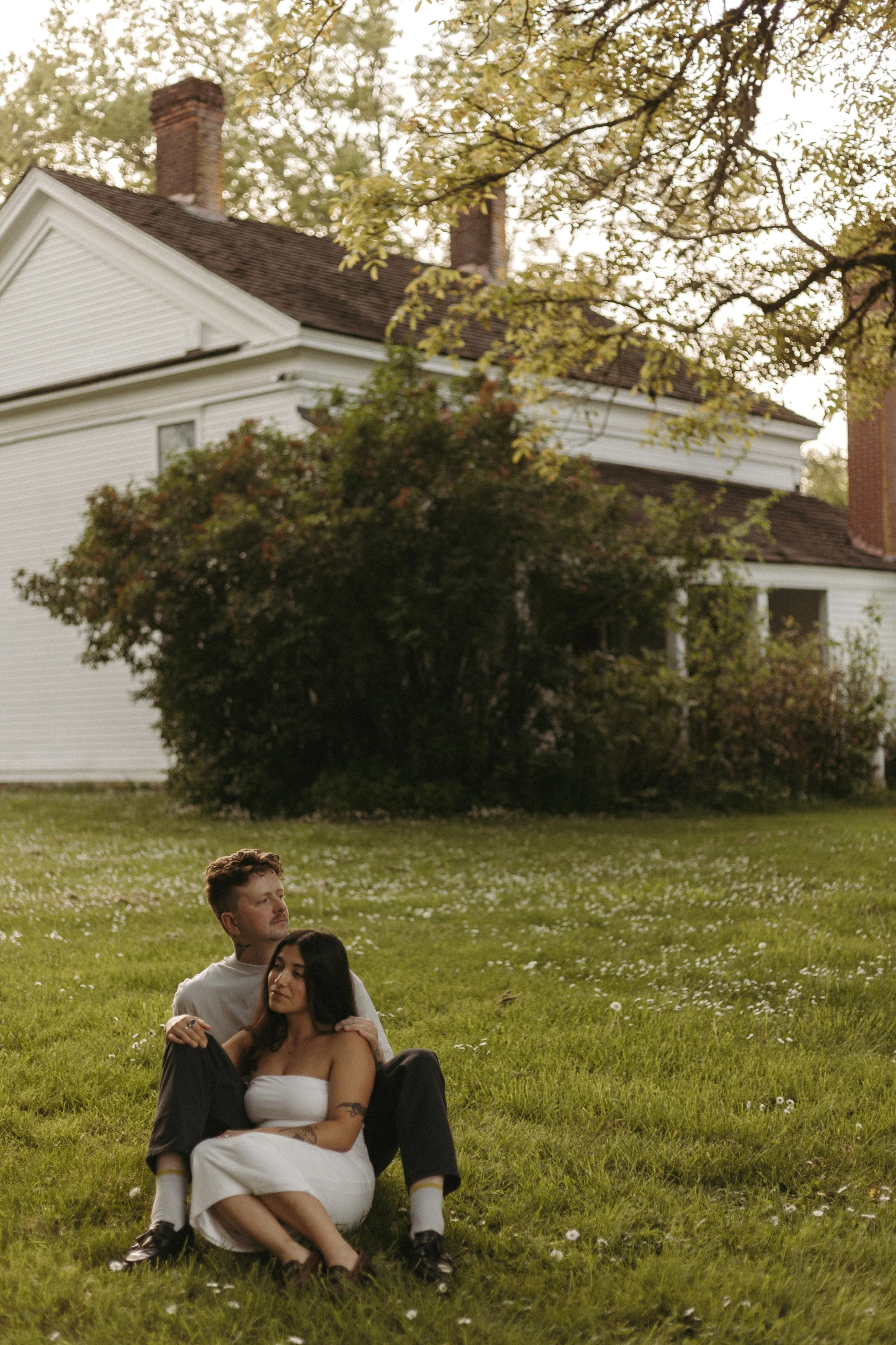 A young couple sitting on the grass in a park with large trees and a white house with a brick chimney in the background during daytime.