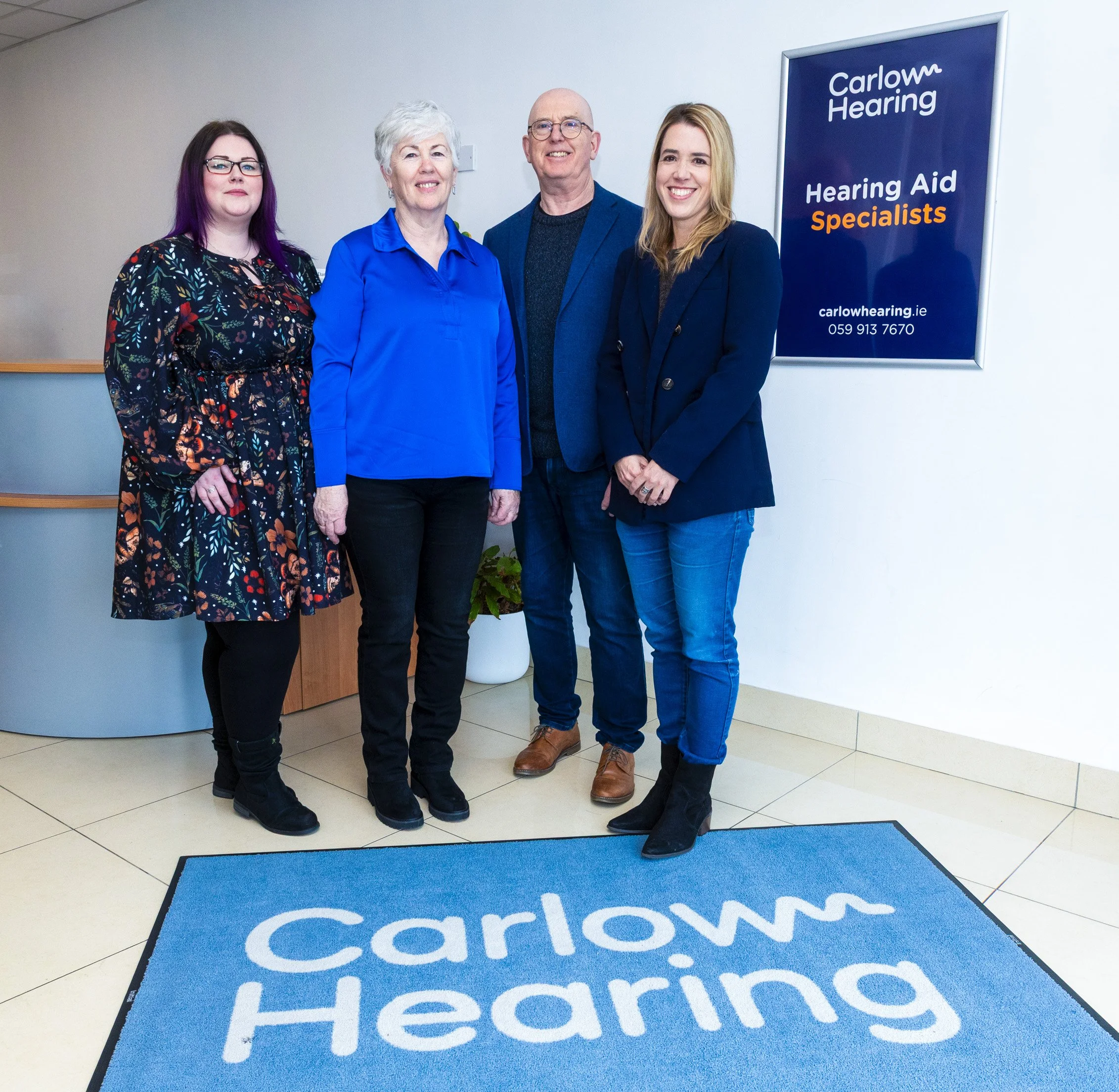 Four people standing inside a building next to a sign that reads 'Carlow Hearing, Hearing Aid Specialists' with the website carlowhearing.ie and contact number 059 913 7670. The group includes three women and one man, all smiling, with a blue mat on the floor that has the word 'Carlow' repeated on it.