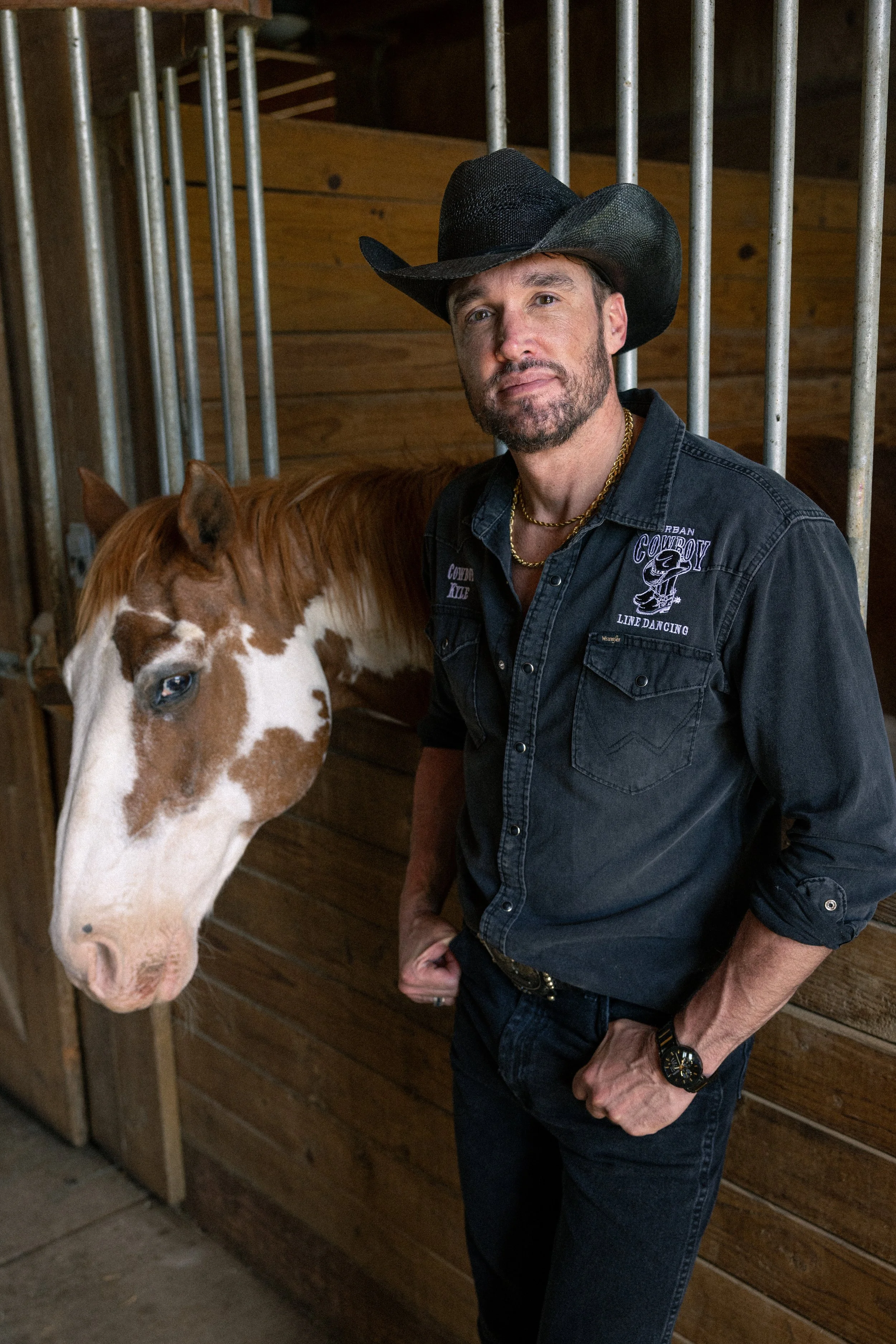 A man dressed in a cowboy hat, denim shirt, and gold jewelry stands next to a brown and white horse inside a wooden stable.
