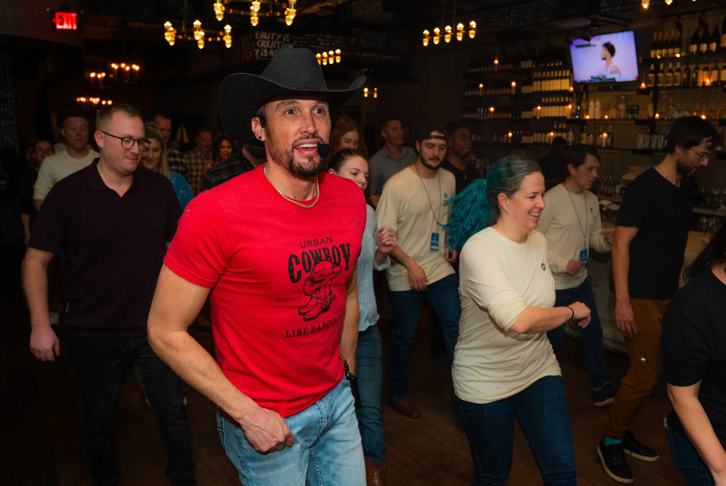 Group of people dancing at a bar or venue, with a man in the foreground wearing a cowboy hat, red t-shirt, and jean shorts, smiling and enjoying the moment.