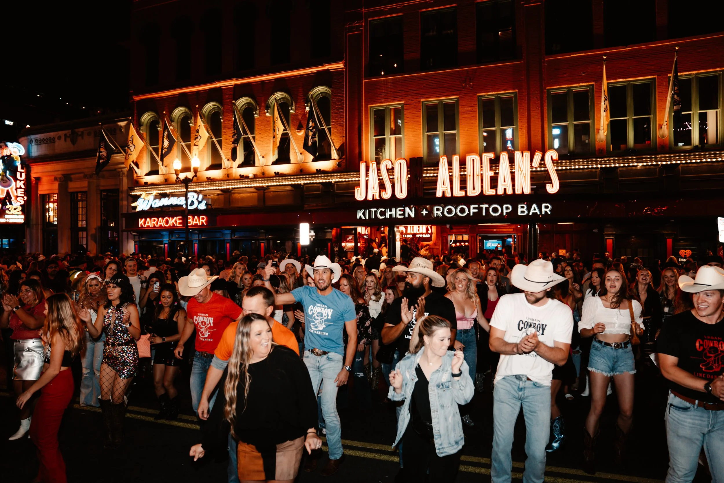 Night scene in an urban area showing a crowded street with many people, some wearing cowboy hats, in front of a brick building illuminated with neon signs. The signs read 'Jasco Aldean's Kitchen + Rooftop Bar' and 'Welcome B2 Karaoke Bar', with flags hanging from the building.