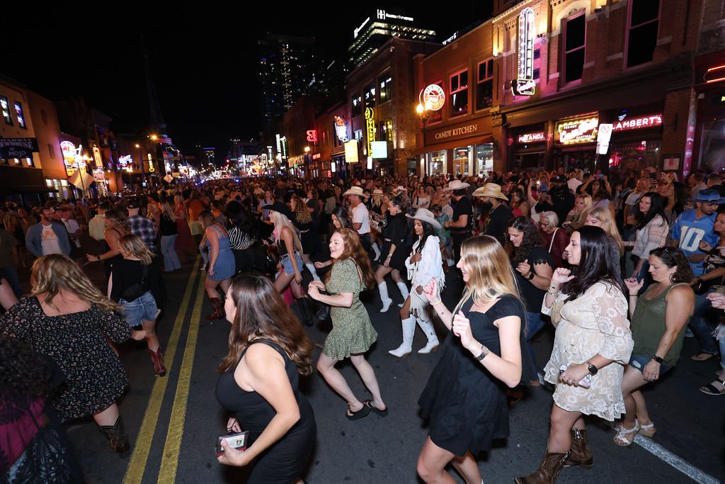 Crowd of people dancing on a city street at night, with neon signs and illuminated storefronts.