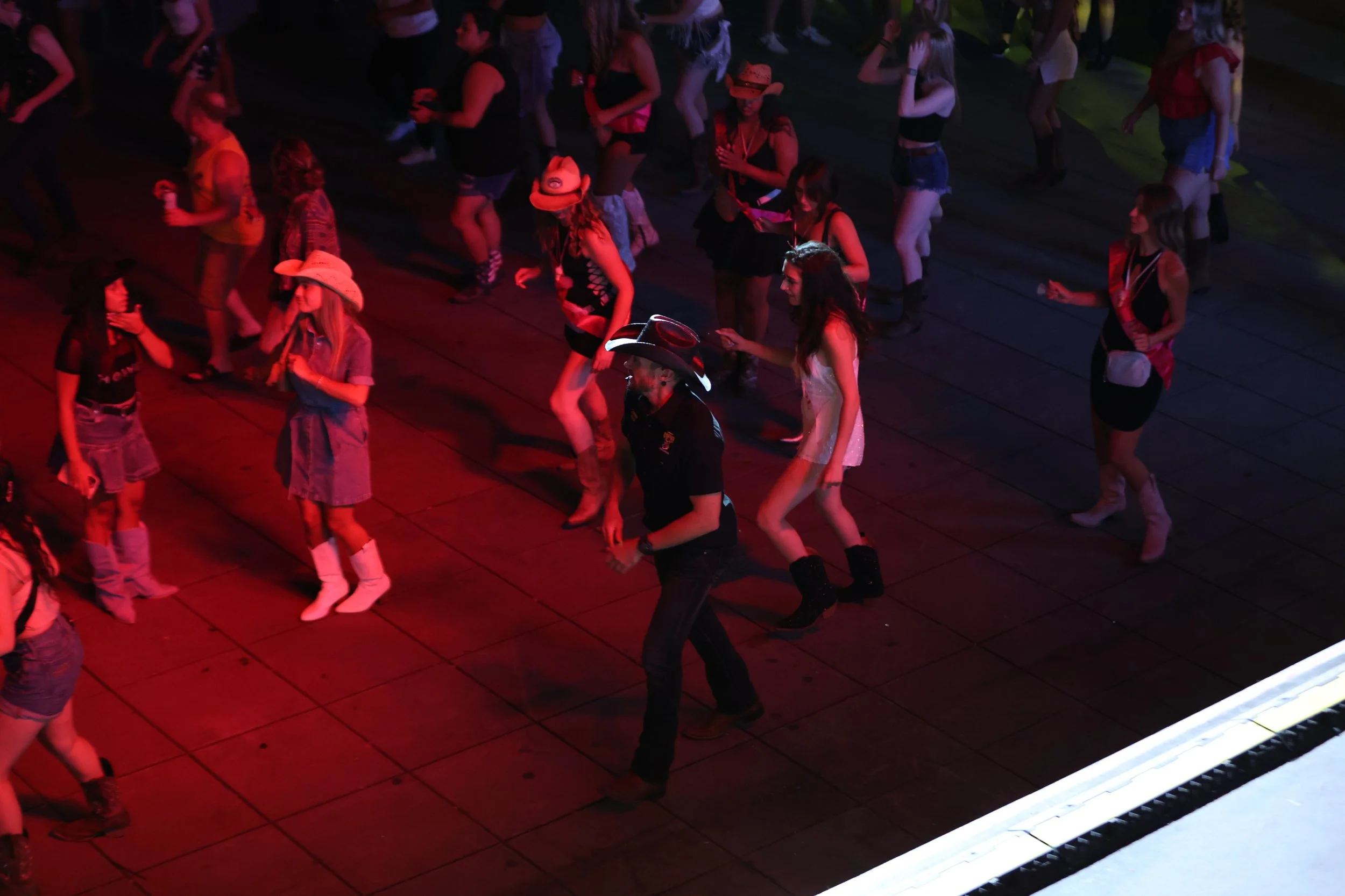 People dancing at a country-themed party or event, with some wearing cowboy hats, plaid shirts, and western-style boots, illuminated by red lighting.